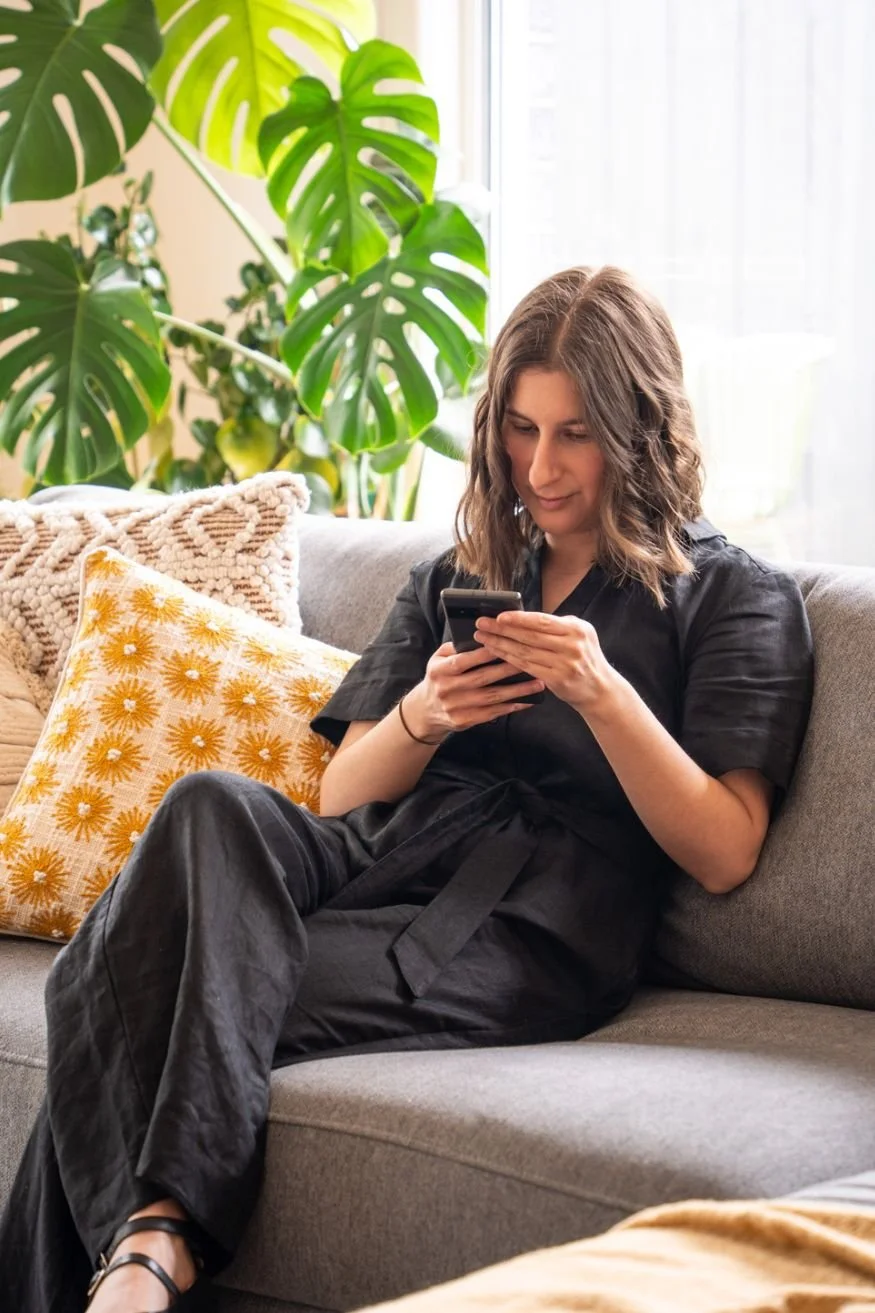 Sarah dressed in black sitting on a comfortable couch with yellow cushions reading copy on her smartphone surrounded by green houseplants