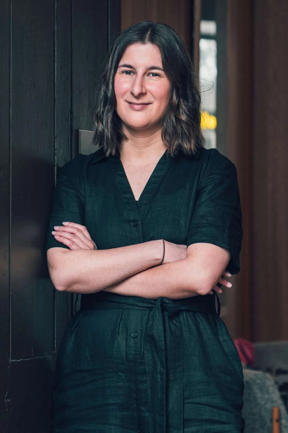 Sarah, website copywriter, looking confident, smiling at the camera with arms folded. She leans against a wall at a cosy cafe dressed in black clothes