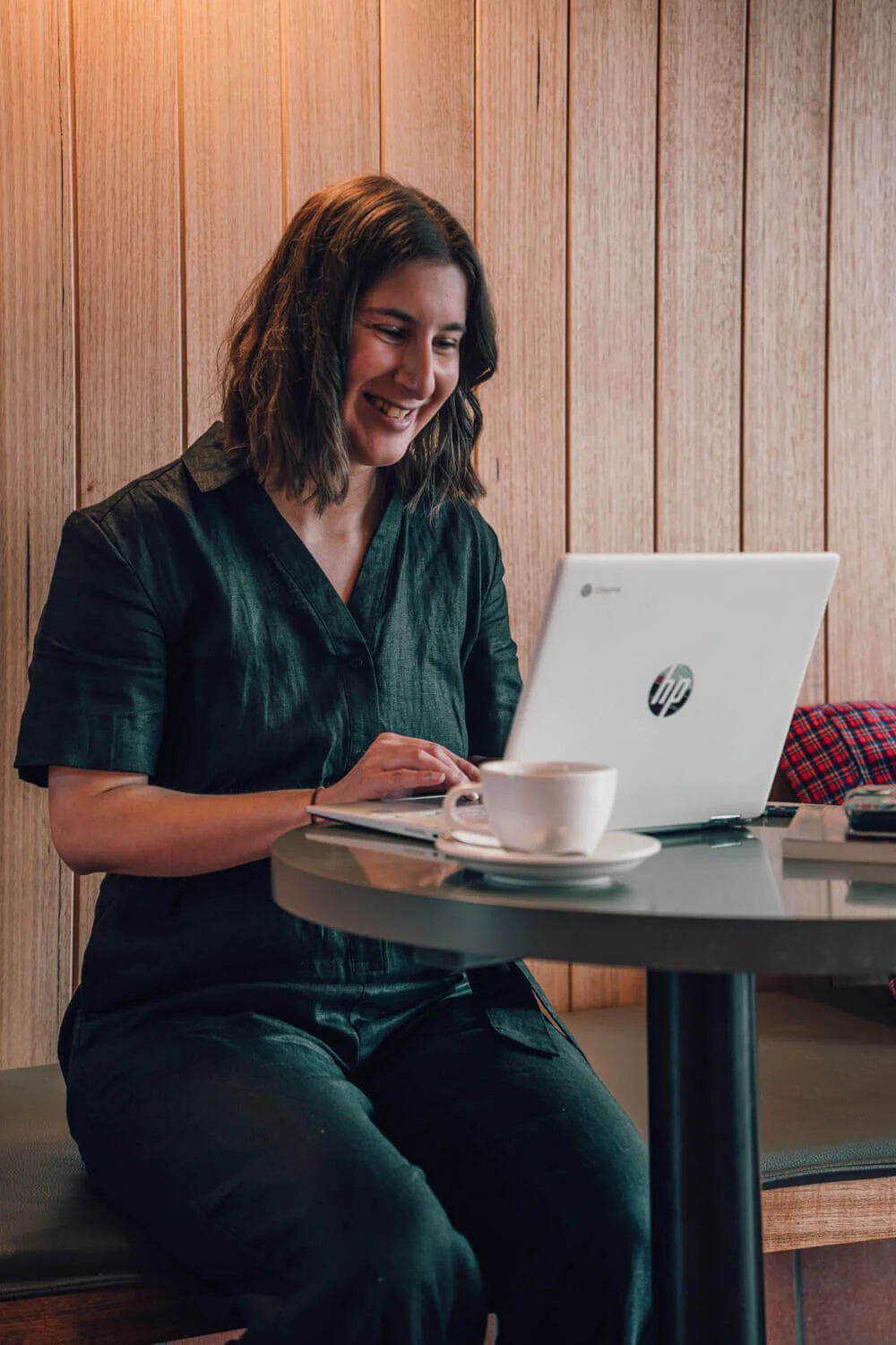 Sarah Marriott smiling in a black outfit working on a laptop, looking at her copywriting portfolio at a café, with a cup of coffee on the table. The setting is cosy and relaxed.