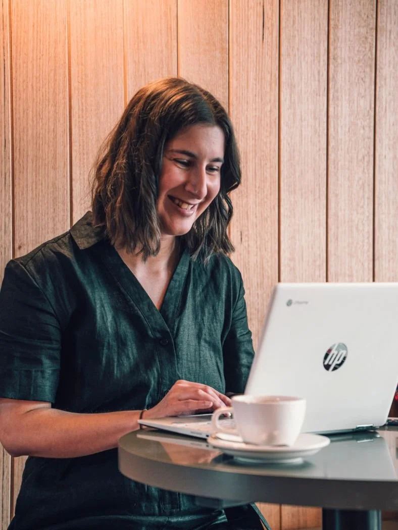 Sarah smiling while working on a laptop at a coffee shop, conveying a sense of joy and engagement in her task