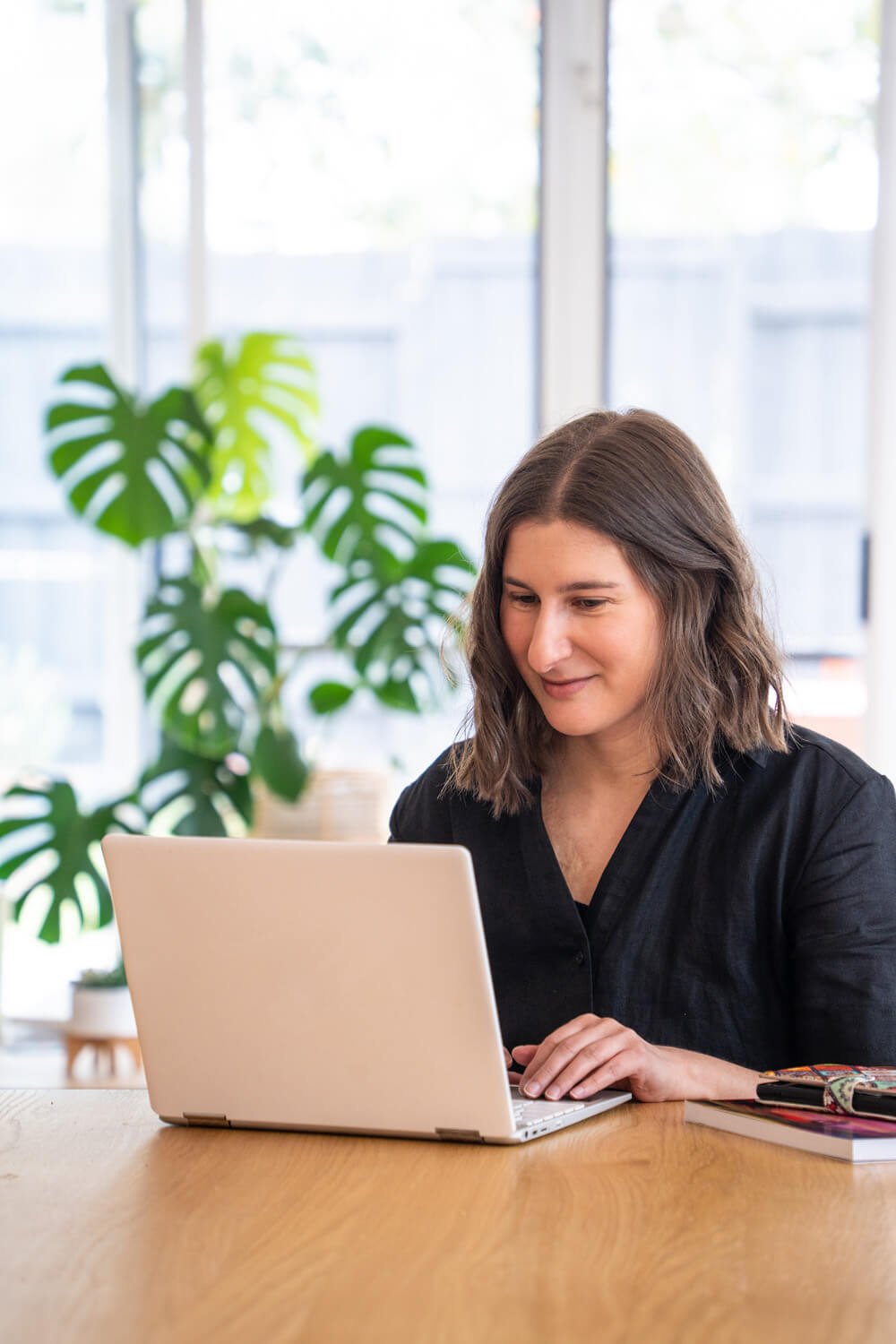 Sarah Marriott with brown hair, wearing black clothes, smiles while working on a laptop at a wooden table. A large green plant is visible in the background.