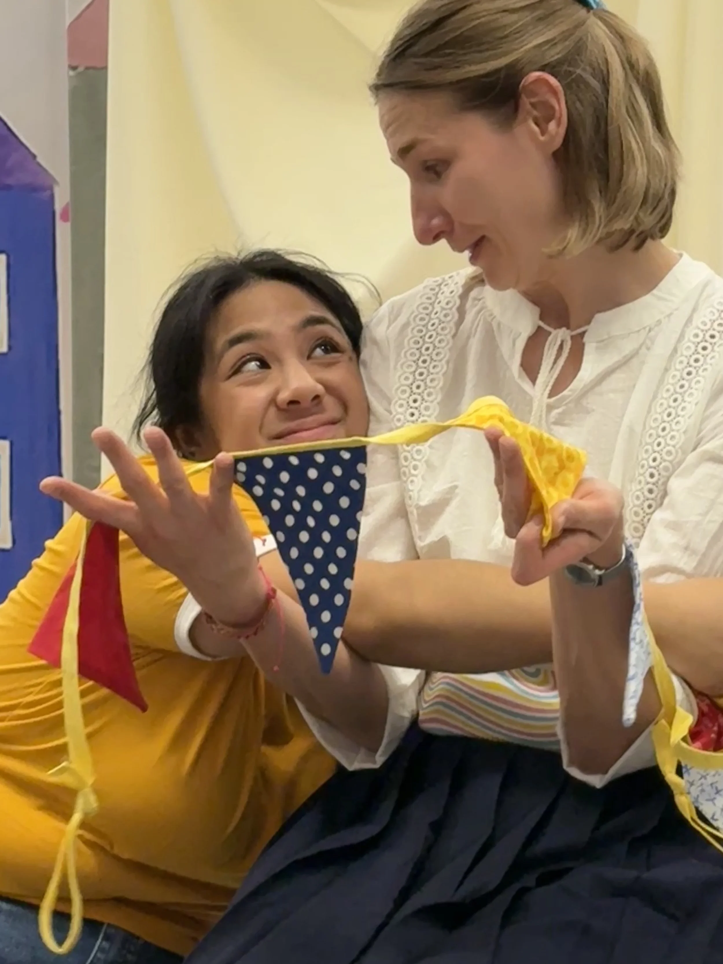 Two women playing little girls, one hugging the other and one showing her a string of flags.