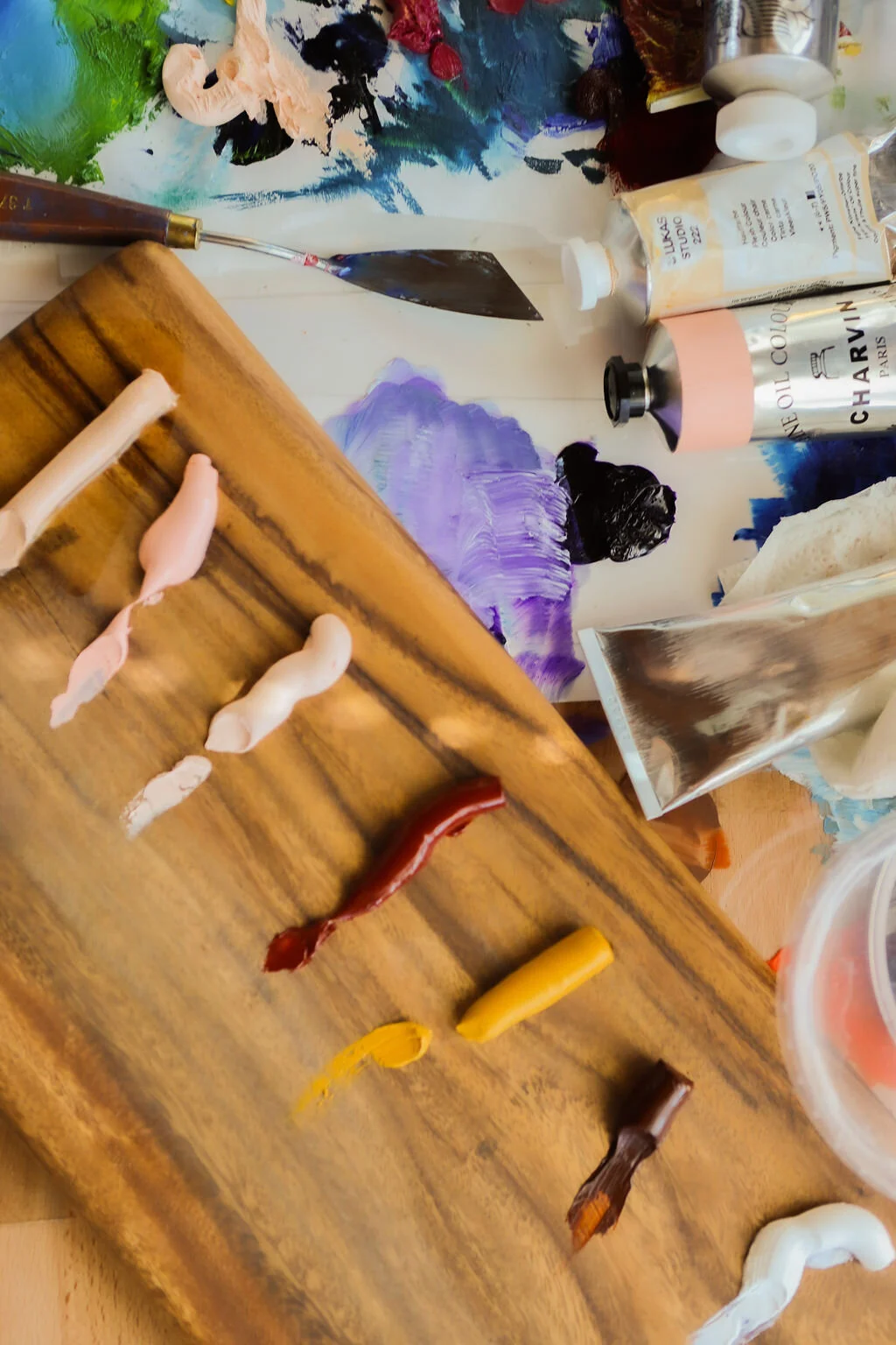A wooden palette with various dollops of paint in different colors including white, red, yellow, and brown, surrounded by artist tools and supplies, including tubes of paint, a palette knife, and a piece of reflective metal.