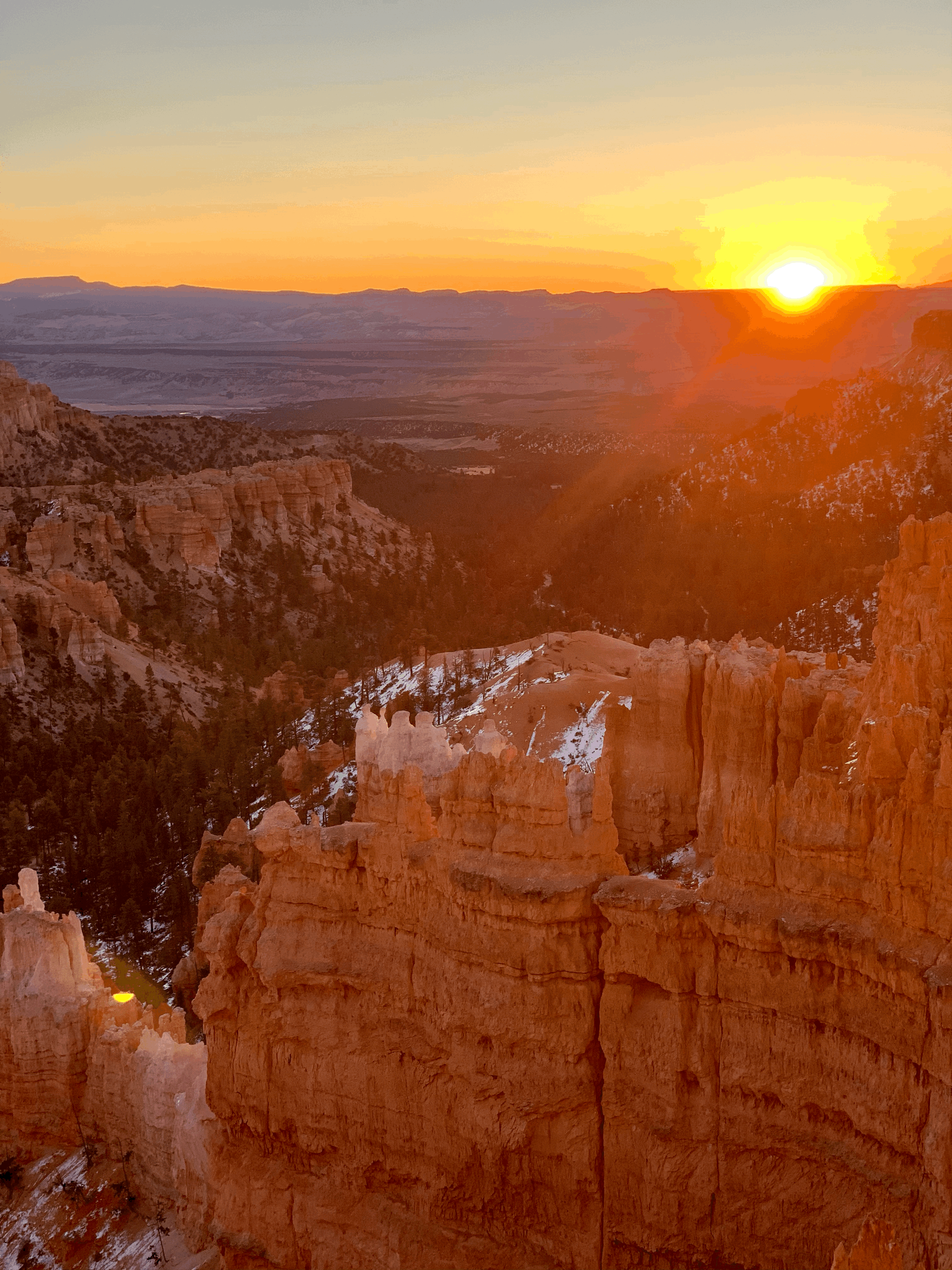Sun setting over Bryce Canyon with orange rock formations and snow patches, distant mountain range under a clear sky.