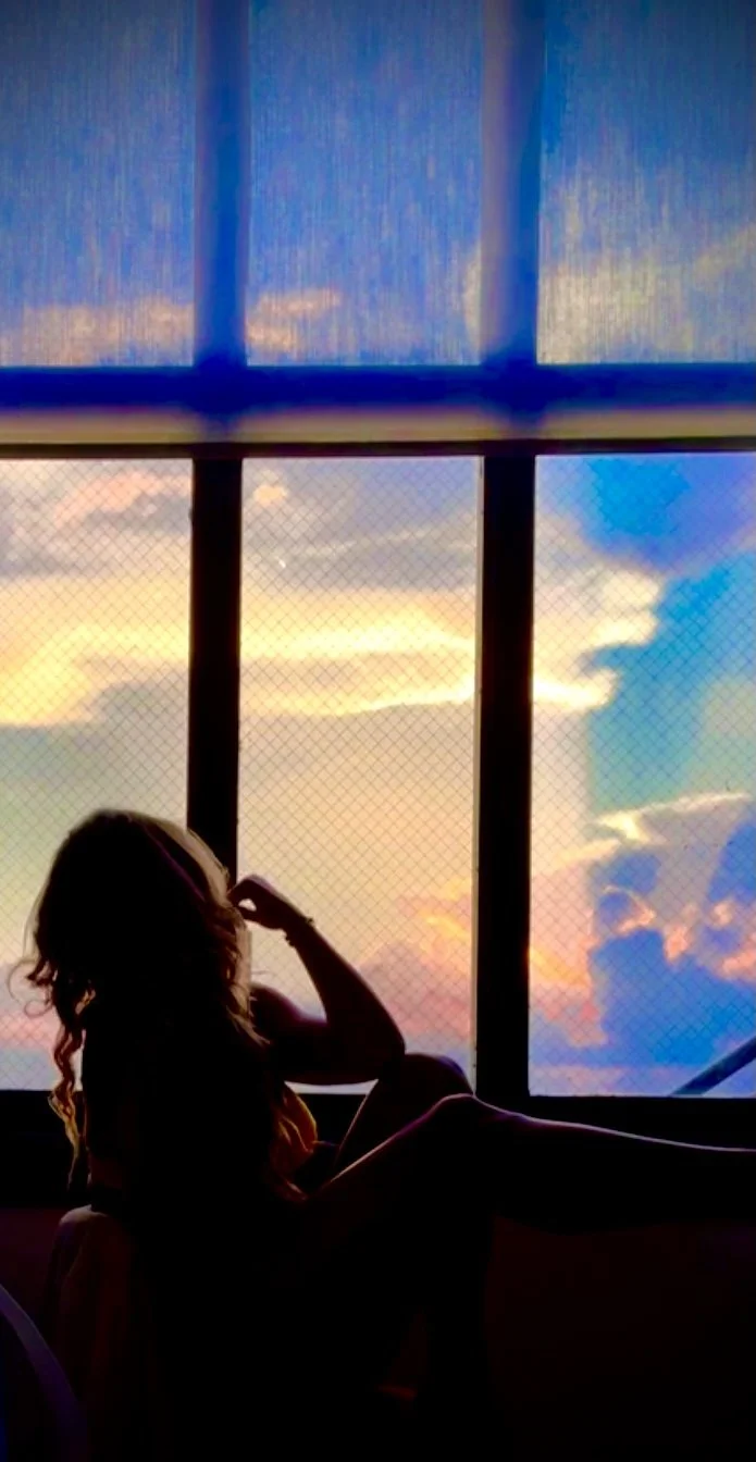 Silhouette of a woman sitting by a window, gazing at a colorful sunset sky with clouds.