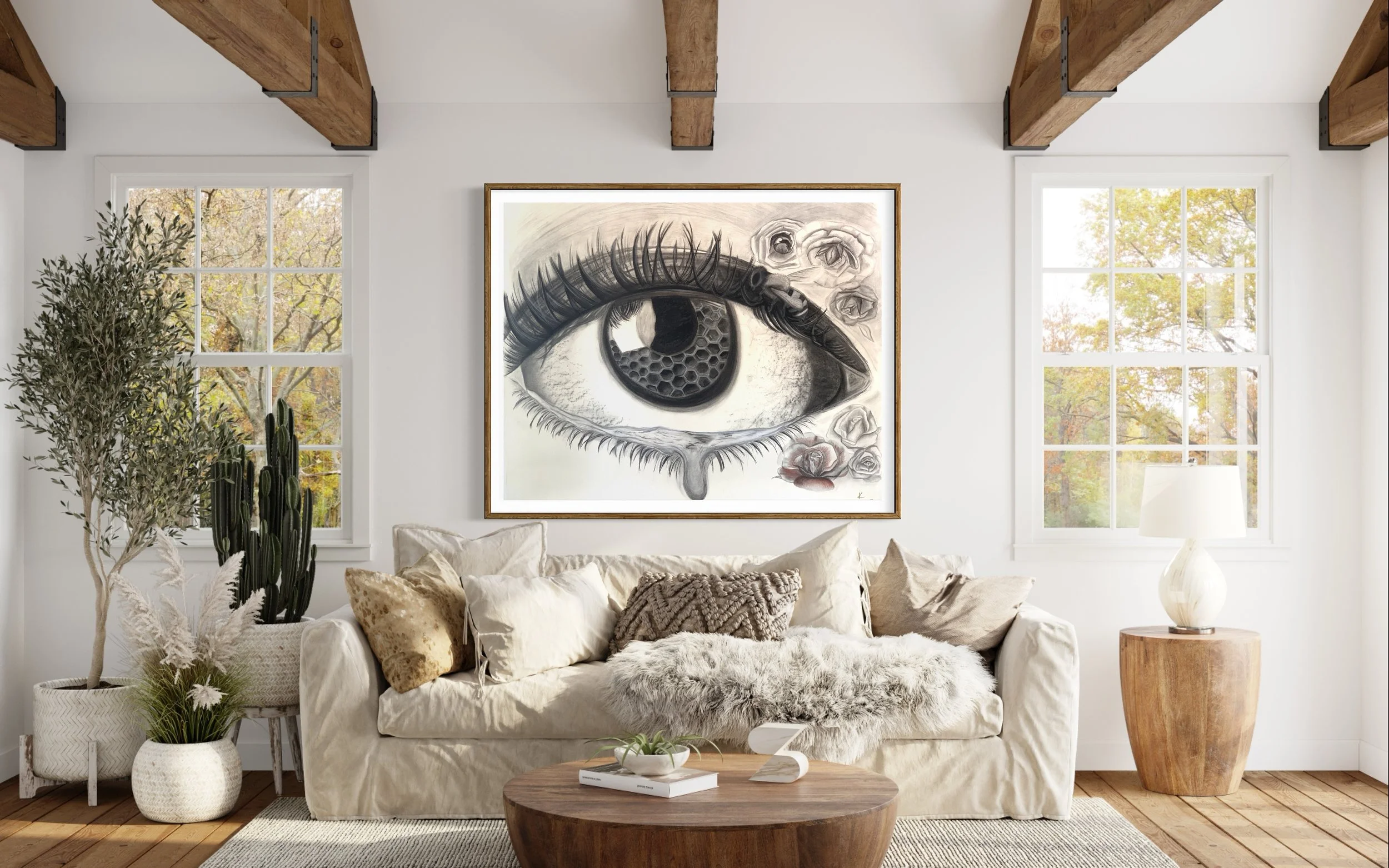 Living room with a large eye painting on the wall, cream sofa with pillows, wooden coffee table, potted plants, window views of trees, and a round wooden side table with a lamp.