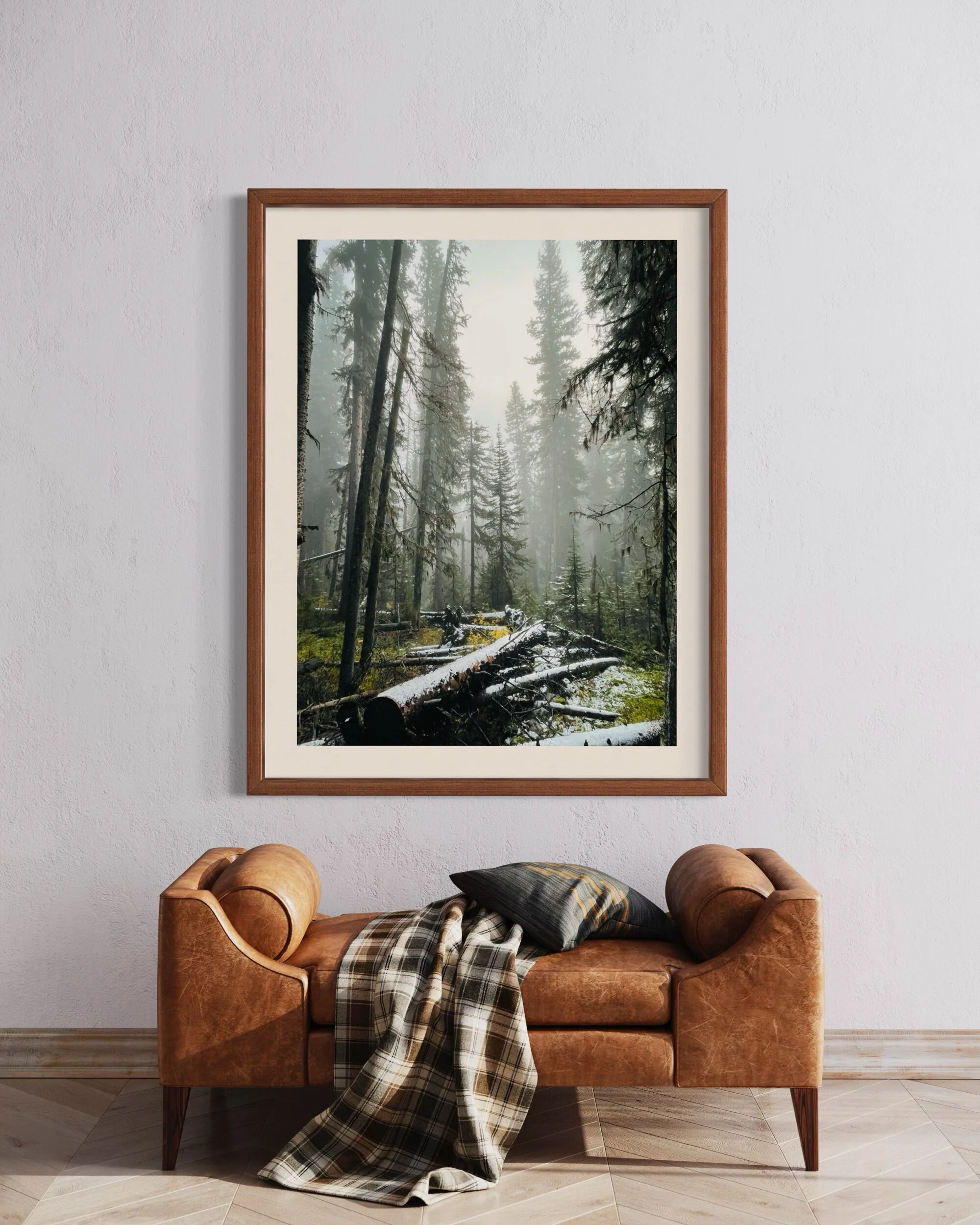 A framed photo of a misty forest hangs on a white wall above a brown leather loveseat with a plaid blanket and black pillow, placed on a beige tiled floor.