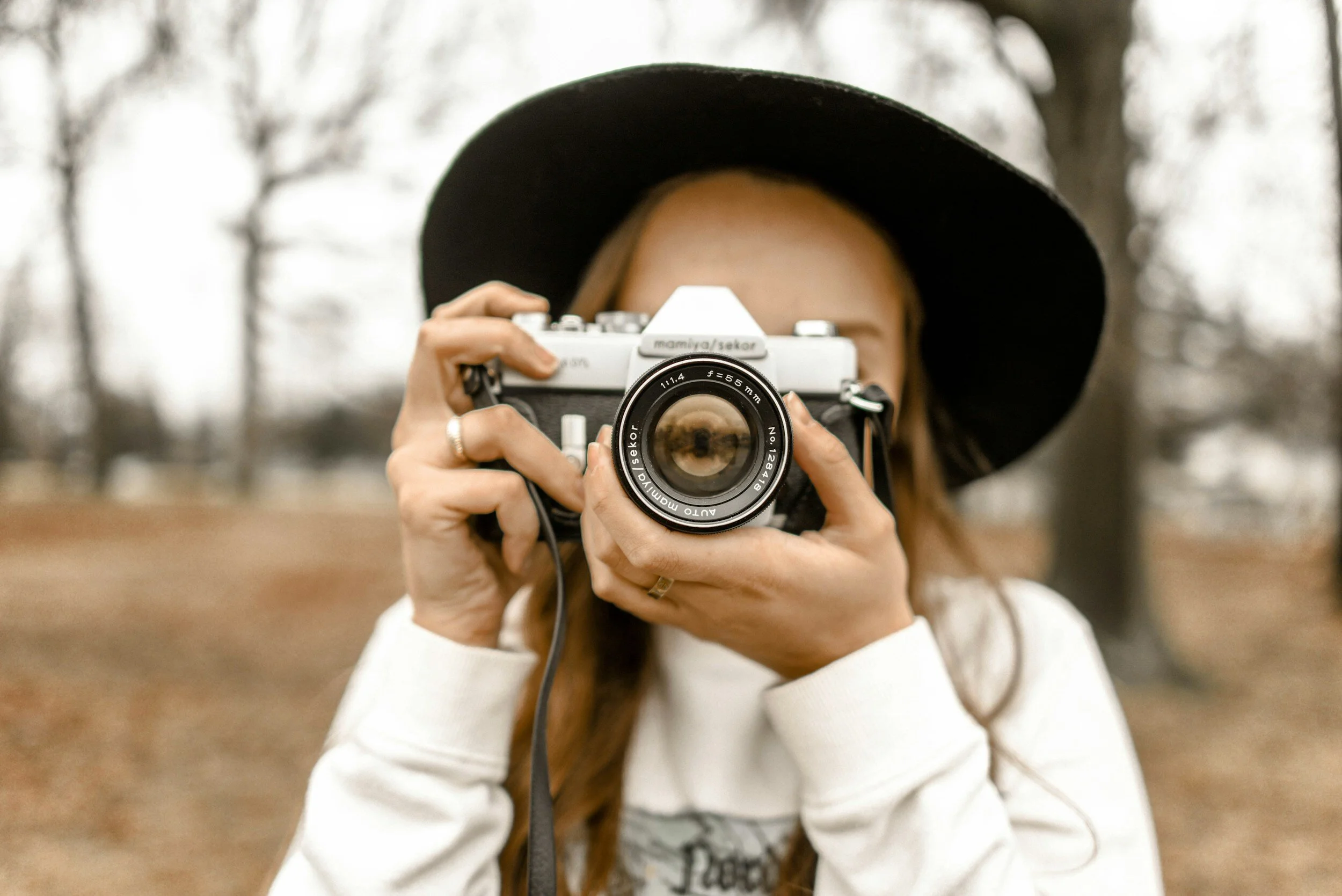 Woman in a black hat taking a photo with a vintage camera outdoors with trees in the background.