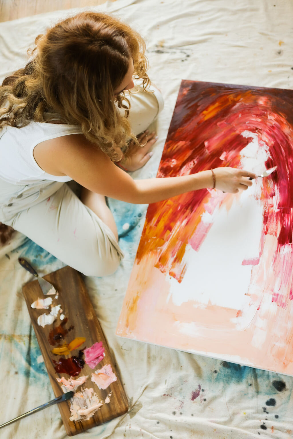A woman with long, curly hair creates an abstract painting on a large canvas, using warm colors like red, orange, and pink.