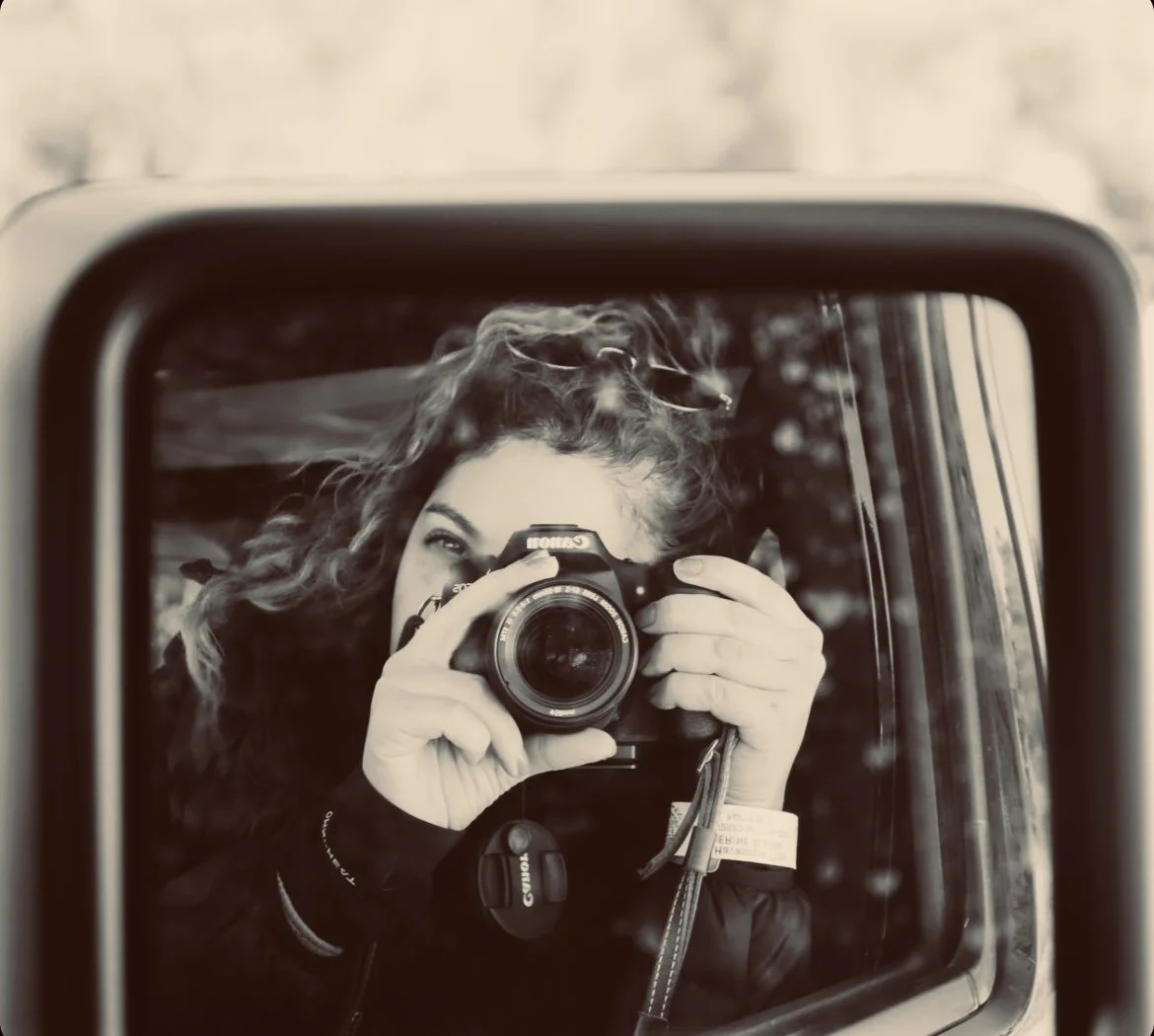 A woman with curly hair taking a selfie in a car's side mirror, holding a Canon camera up to her face.