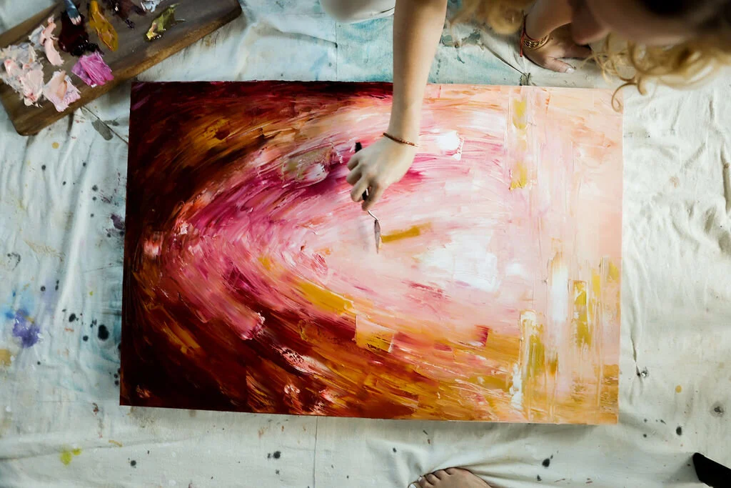 Close-up of a person painting an abstract artwork with pink, white, and yellow brushstrokes on a large canvas, placed on a white paper-covered floor with paint splatters.