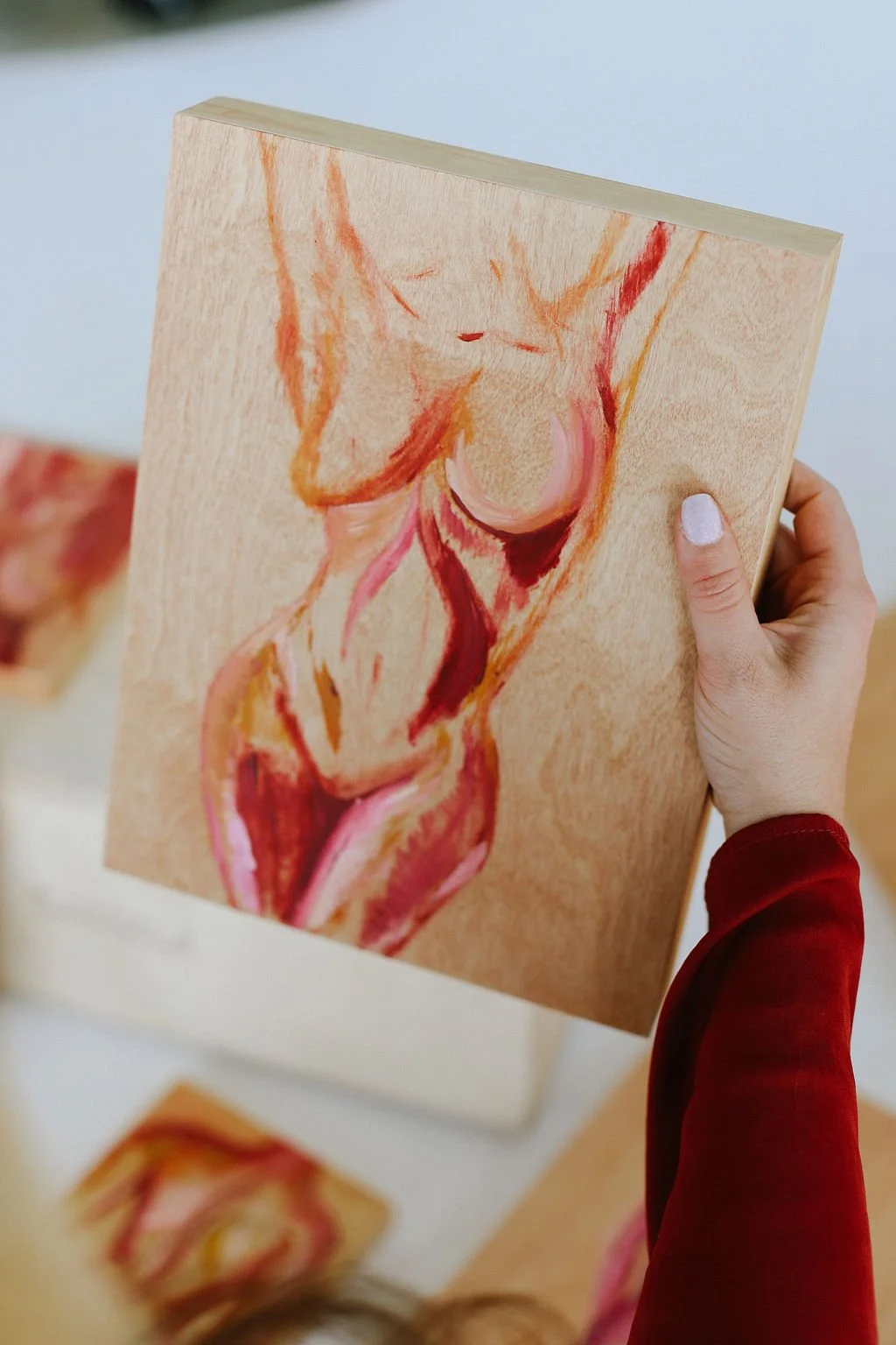 A person holding a wooden painting of a figurative painted female, with additional similar paintings in the background.