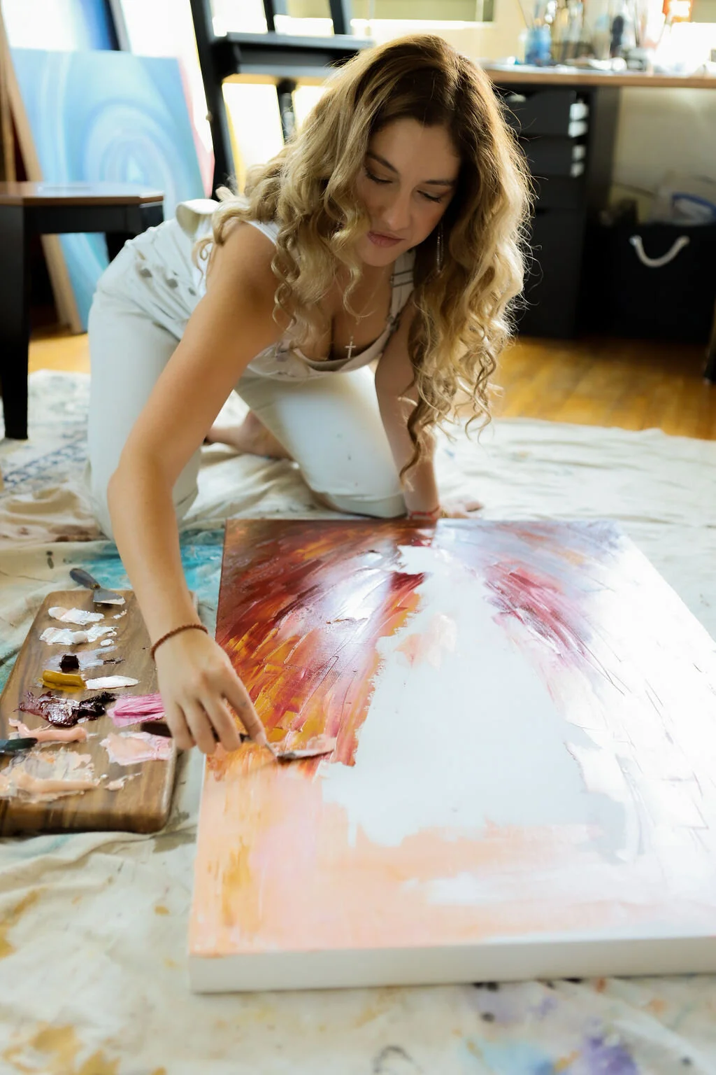 A woman with long curly hair is painting on a large canvas on the floor, using a palette with different colors of paint nearby.