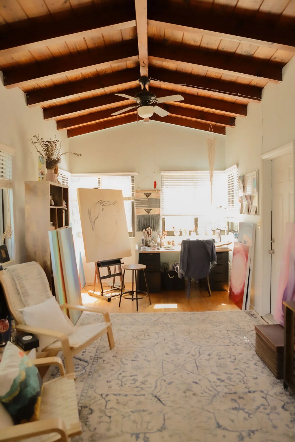 A cozy artist's workspace with a wooden vaulted ceiling, ceiling fan, and natural light from multiple windows. There is an easel with a sketch, a desk with art supplies, and various paintings and artwork around the room. The space includes a beige ch