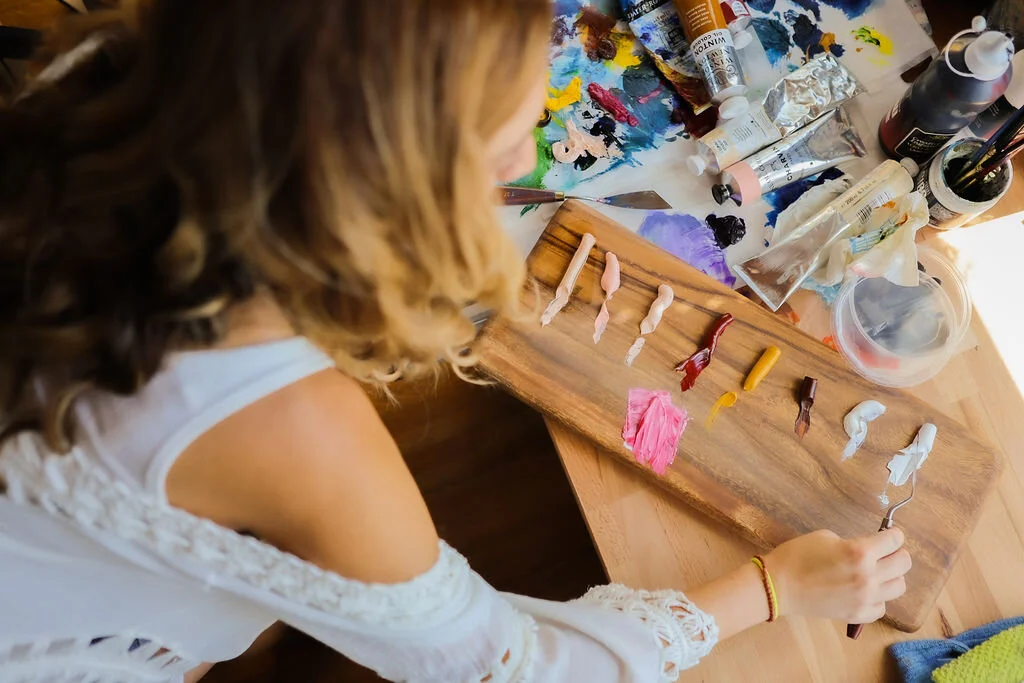 A woman is painting colored candle wax on a wooden board, with various paint tubes and brushes on a colorful palette and a cluttered table nearby.