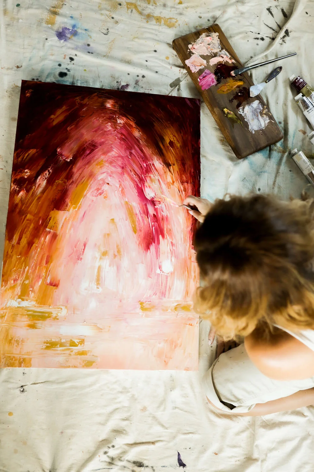 A person with brown hair painting an abstract artwork on a large canvas, using a variety of colors including pink, red, white, and gold, with painting supplies and brushes nearby on a paint-splattered cloth.