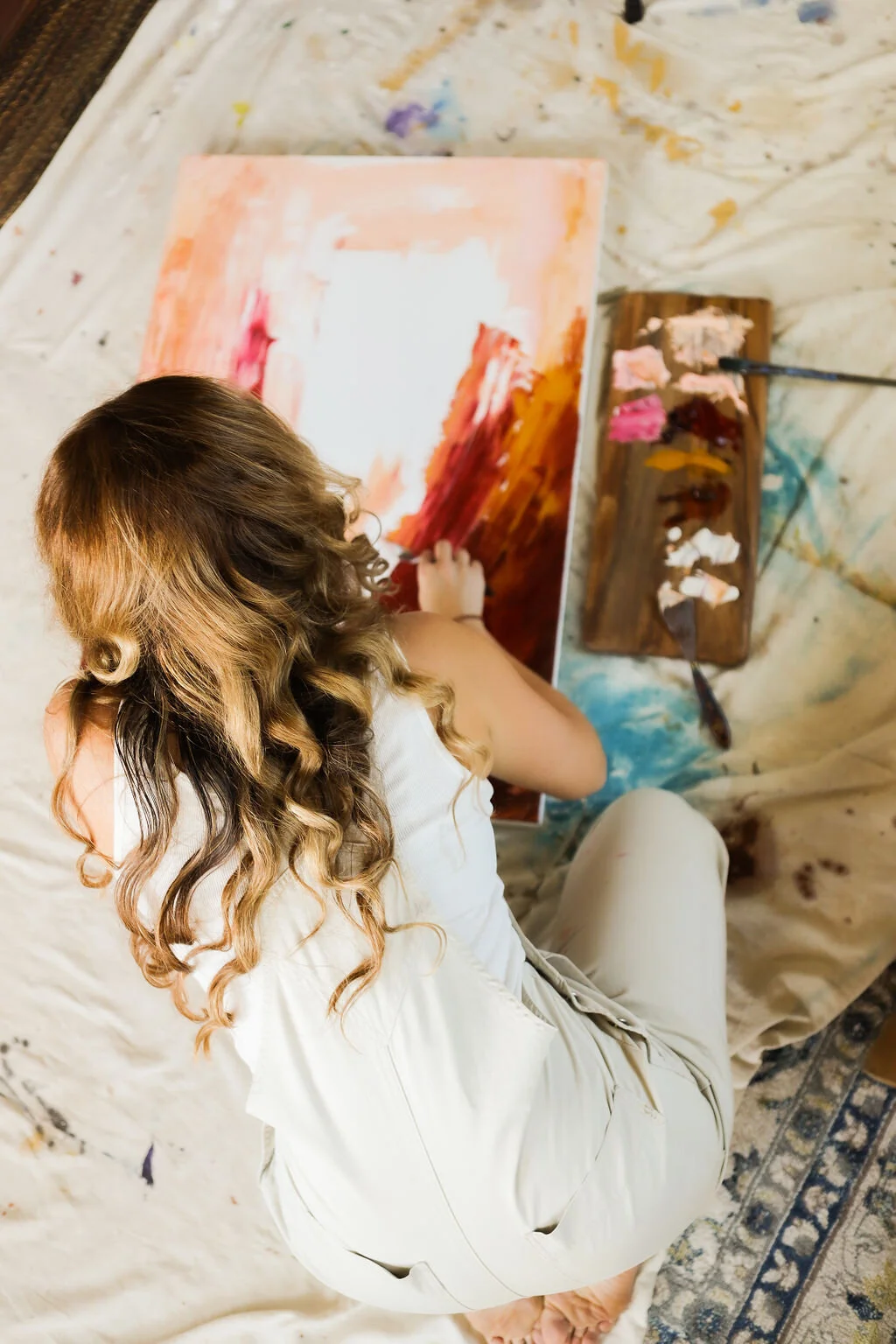 A woman with long, curly hair is sitting on a sheet-covered surface, painting an abstract artwork on a canvas with warm colors, using a palette and brushes placed on a wooden work table.