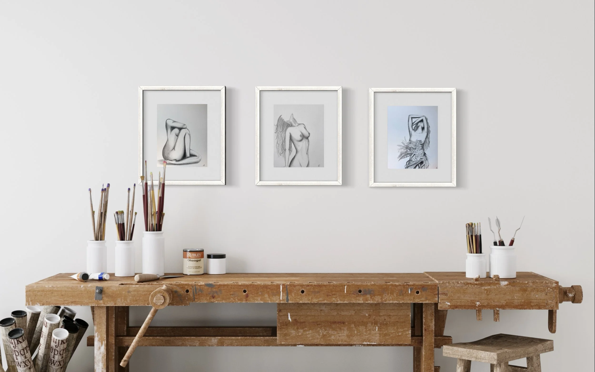 Art studio workspace with a wooden workbench, jars of paintbrushes, and three framed sketches of female figures on the white wall.