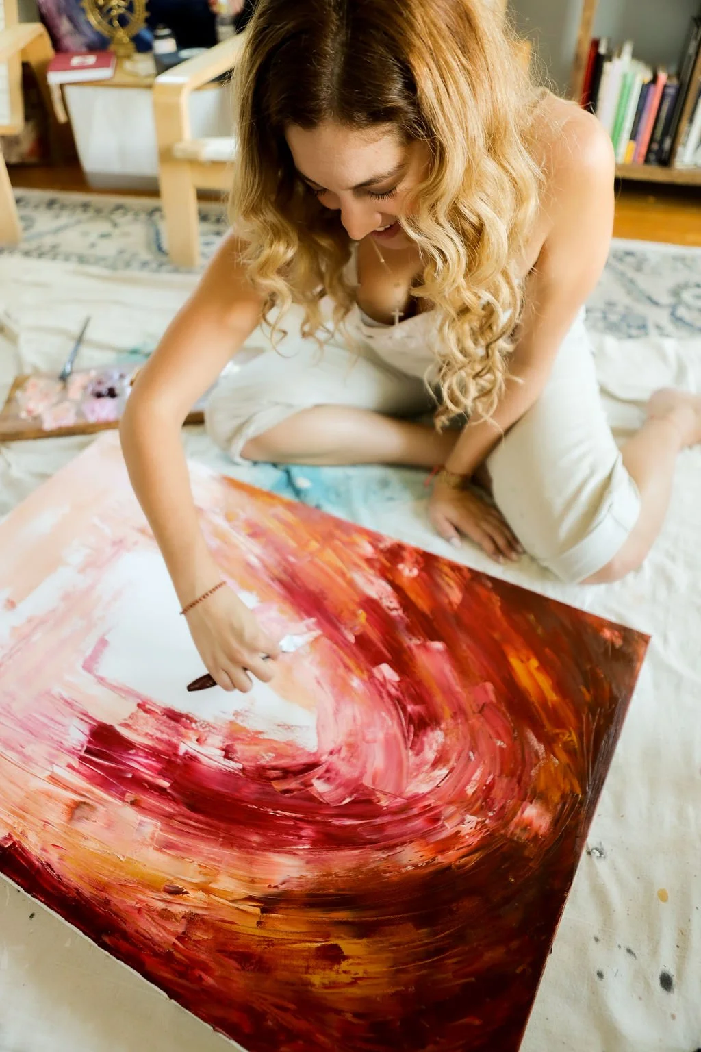 A woman with long curly hair is sitting on the floor, painting an abstract artwork with red, pink, and orange hues on a canvas.