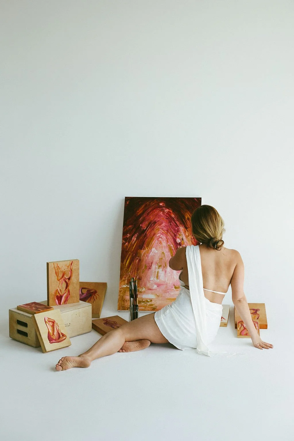 A woman with blonde hair in a low bun, partially wrapped in a white cloth, sitting on a white floor surrounded by abstract paintings and art supplies in a minimalist studio with a plain white wall.