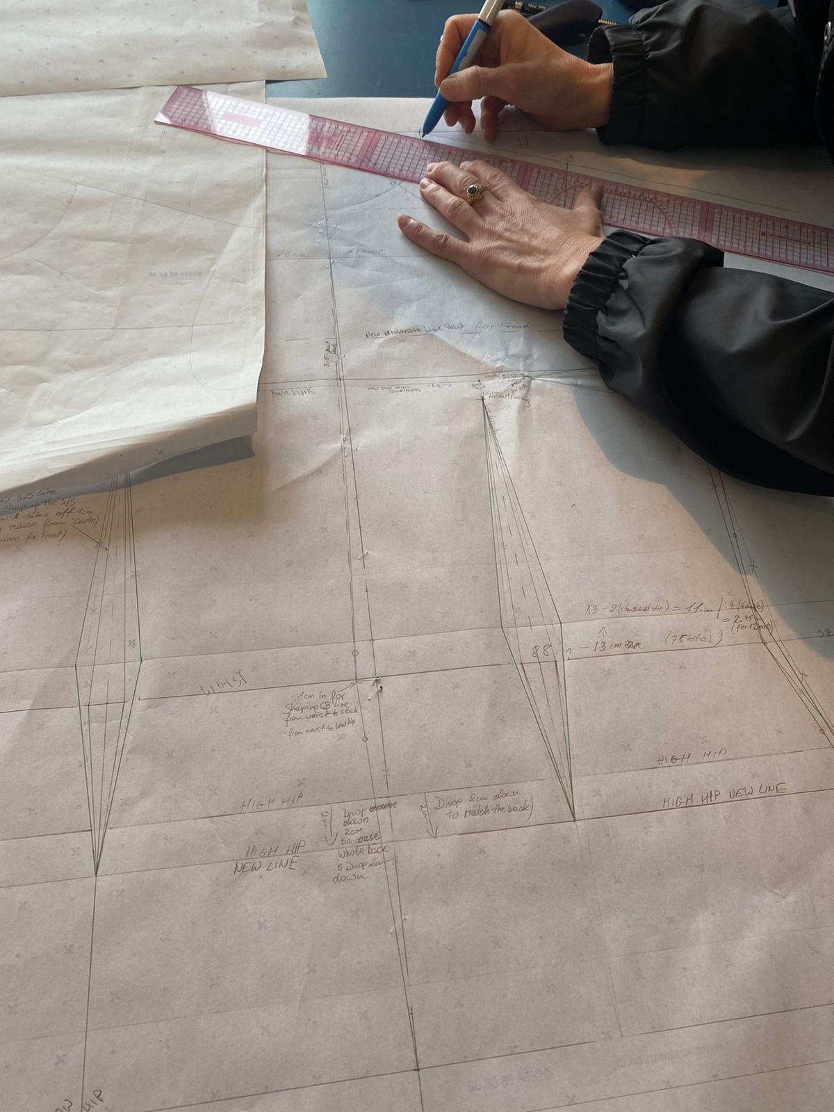 A person is drawing on a large technical or architectural blueprint with a blue pen, using a pink ruler. The blueprint contains detailed sketches and measurements related to high hip design.