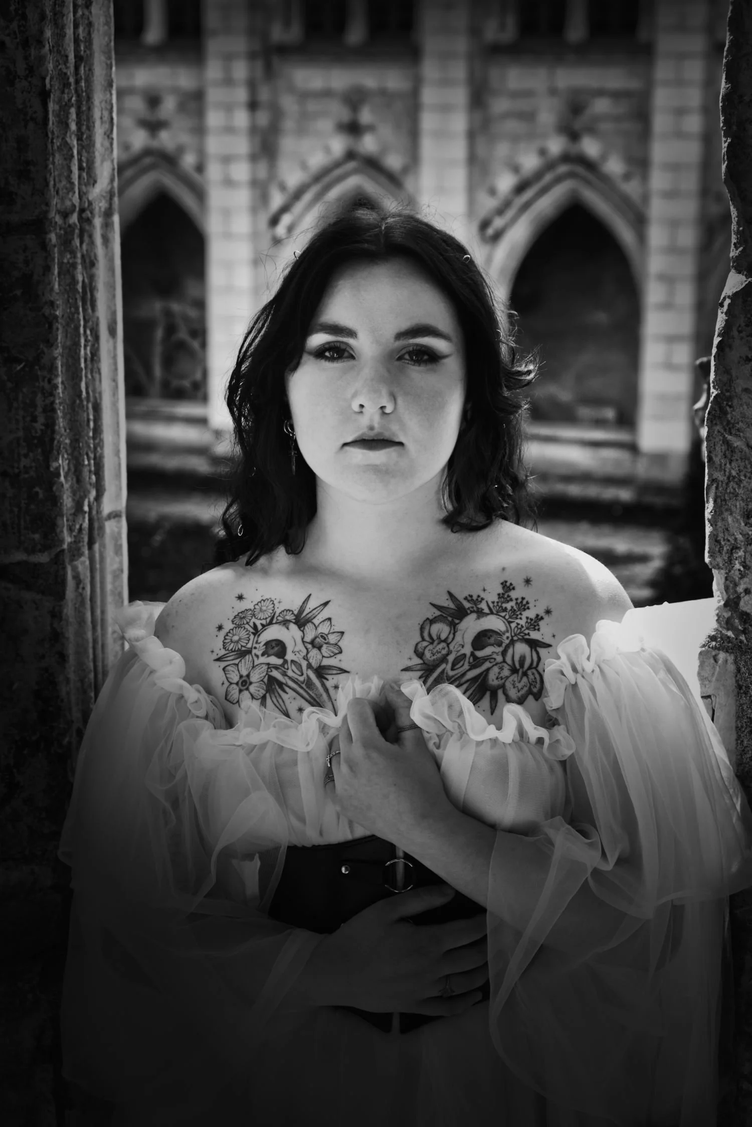 Une femme avec des tatouages de crânes et de fleurs, portant une robe à épaules dénudées, se tient entre deux piliers en pierre devant une structure gothique, dans une ambiance en noir et blanc.