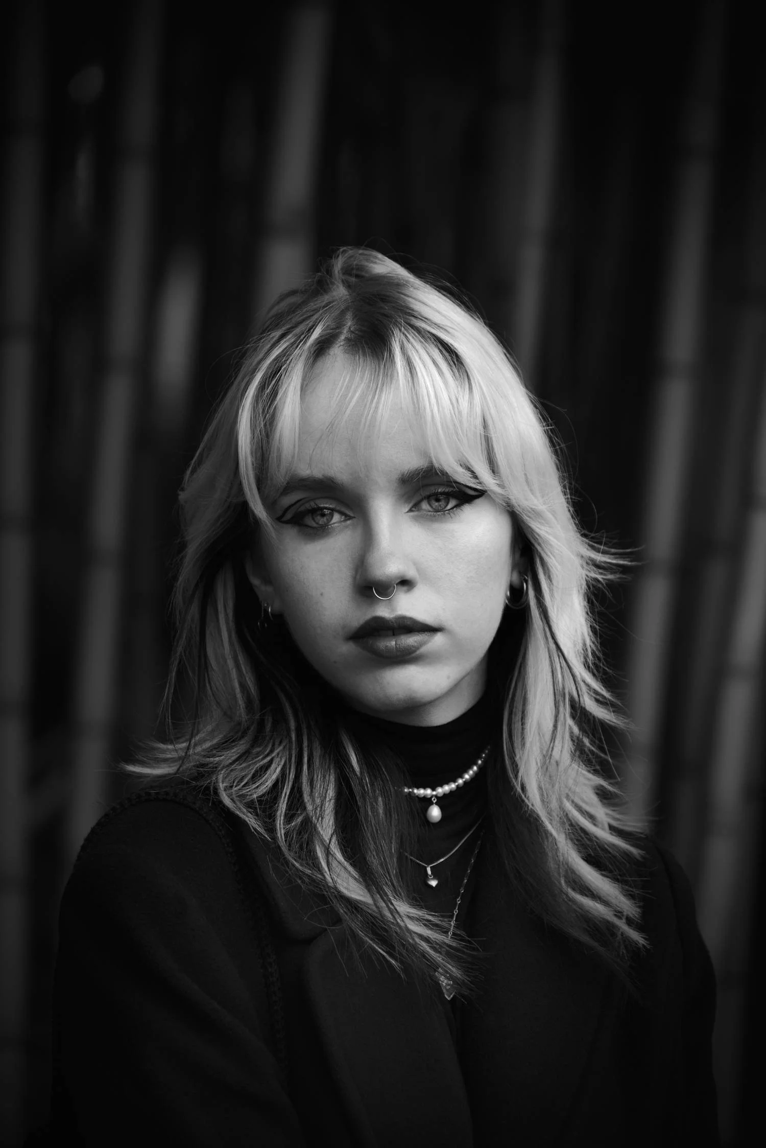 Portrait en noir et blanc d'une jeune femme avec des cheveux blonds, maquillage traditionnel avec du eyeliner, plusieurs colliers et boucles d'oreilles, peignant un regard vers l'objectif, en arrière-plan des bambous.