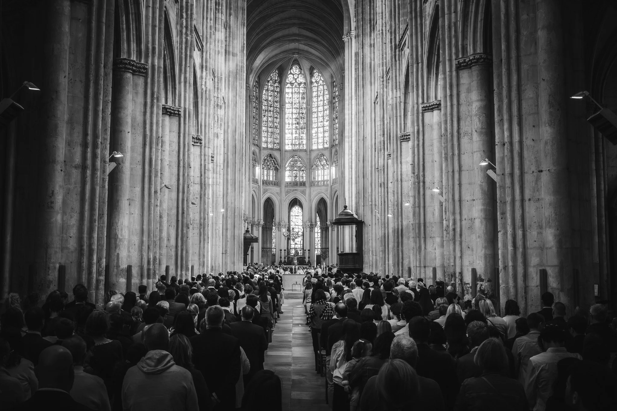 Une foule de personnes dans une cathédrale gothique en pierre, assises ou debout, regardant vers l'autel au fond, avec de hautes fenêtres en vitrail lumineux.
