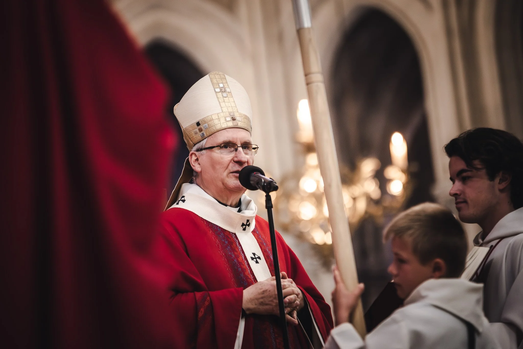 Un évêque en vêtements rouges et or, portant une mitre blanche, parle dans un micro lors d'une cérémonie religieuse dans une église, entouré de jeunes acolytes.