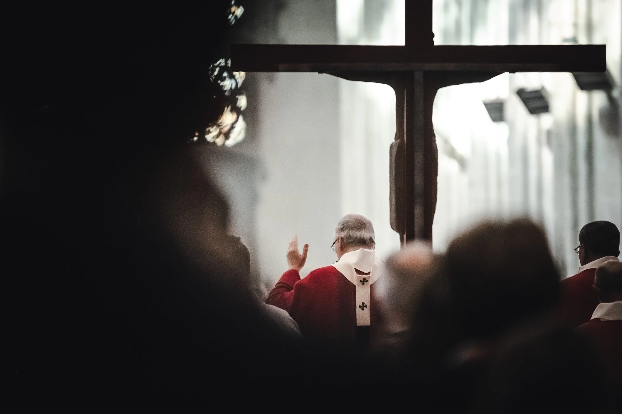 Un prêtre catholique en robe rouge avec une croix blanche au dos, levant la main lors d'une cérémonie religieuse dans une église. Les silhouettes de personnes assistent à la cérémonie, avec des rideaux laissant passer la lumière en arrière-plan.