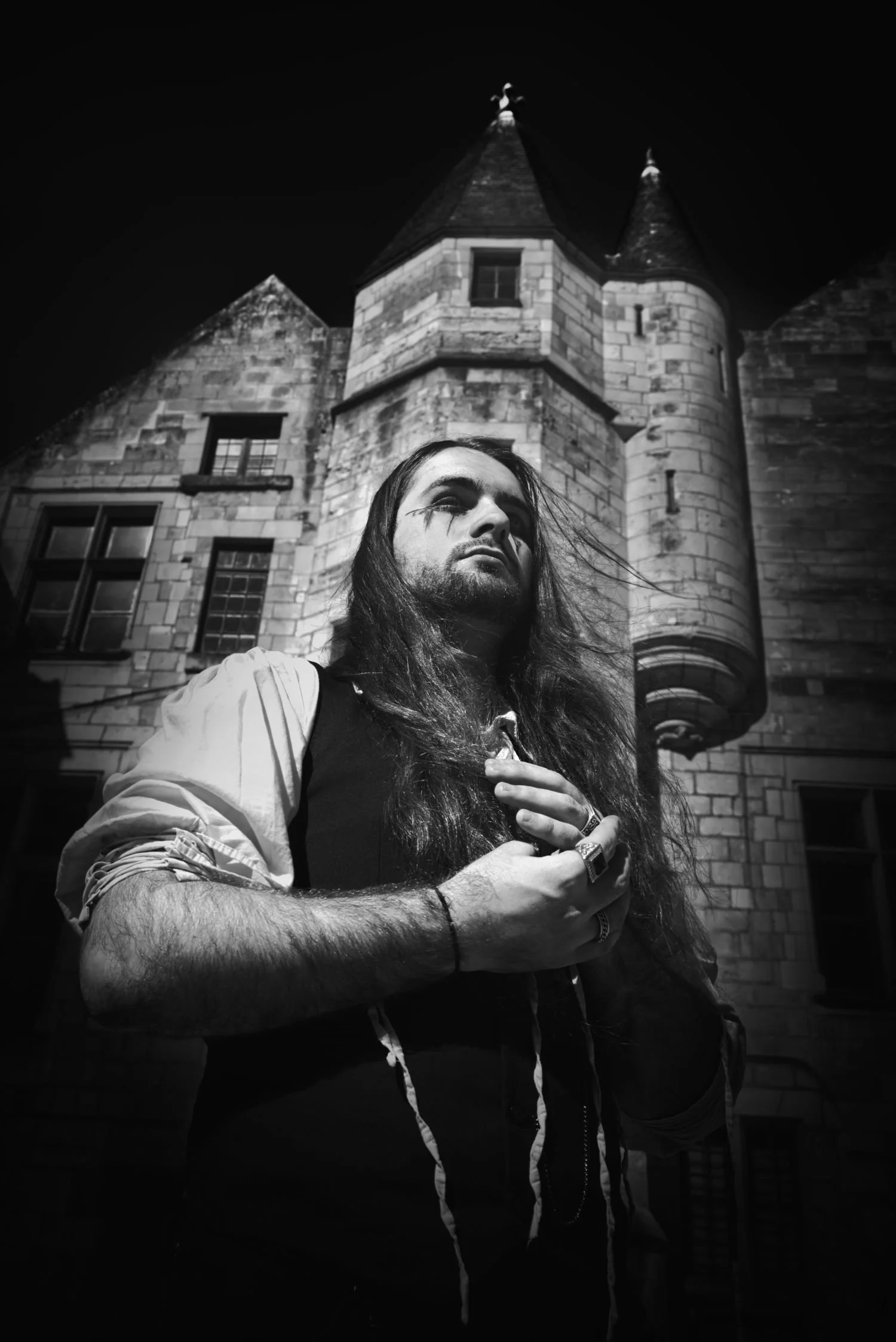Un homme avec de longs cheveux et un maquillage sombre posant devant un château en pierre la nuit.