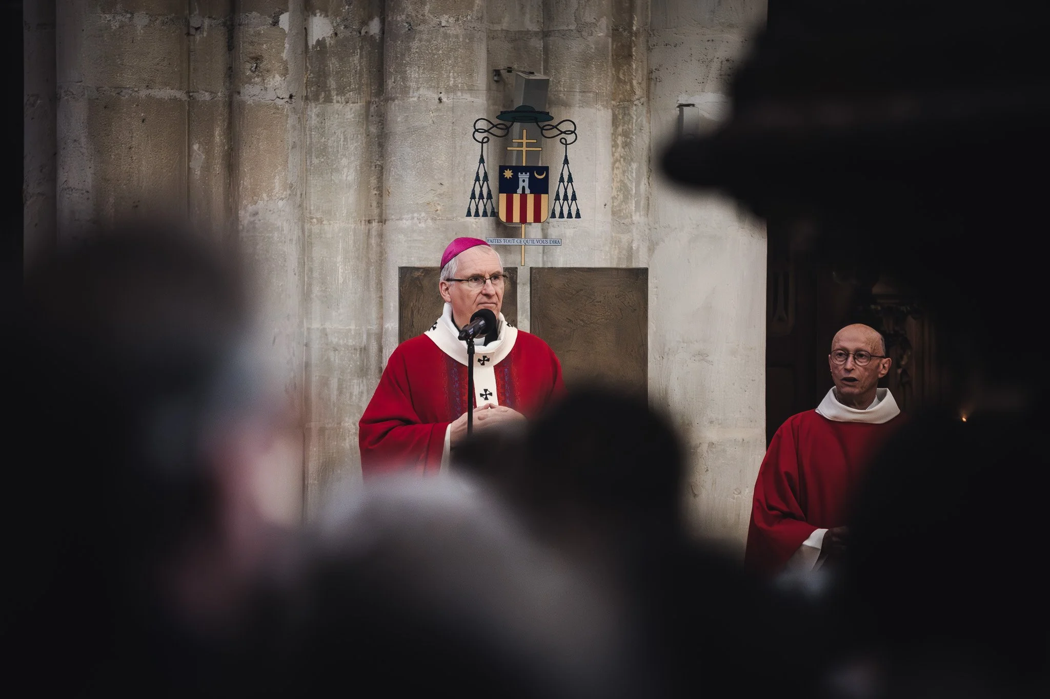 Un évêque en tenue rouge parle dans une église, entouré de personnes, avec un symbole religieux en arrière-plan.