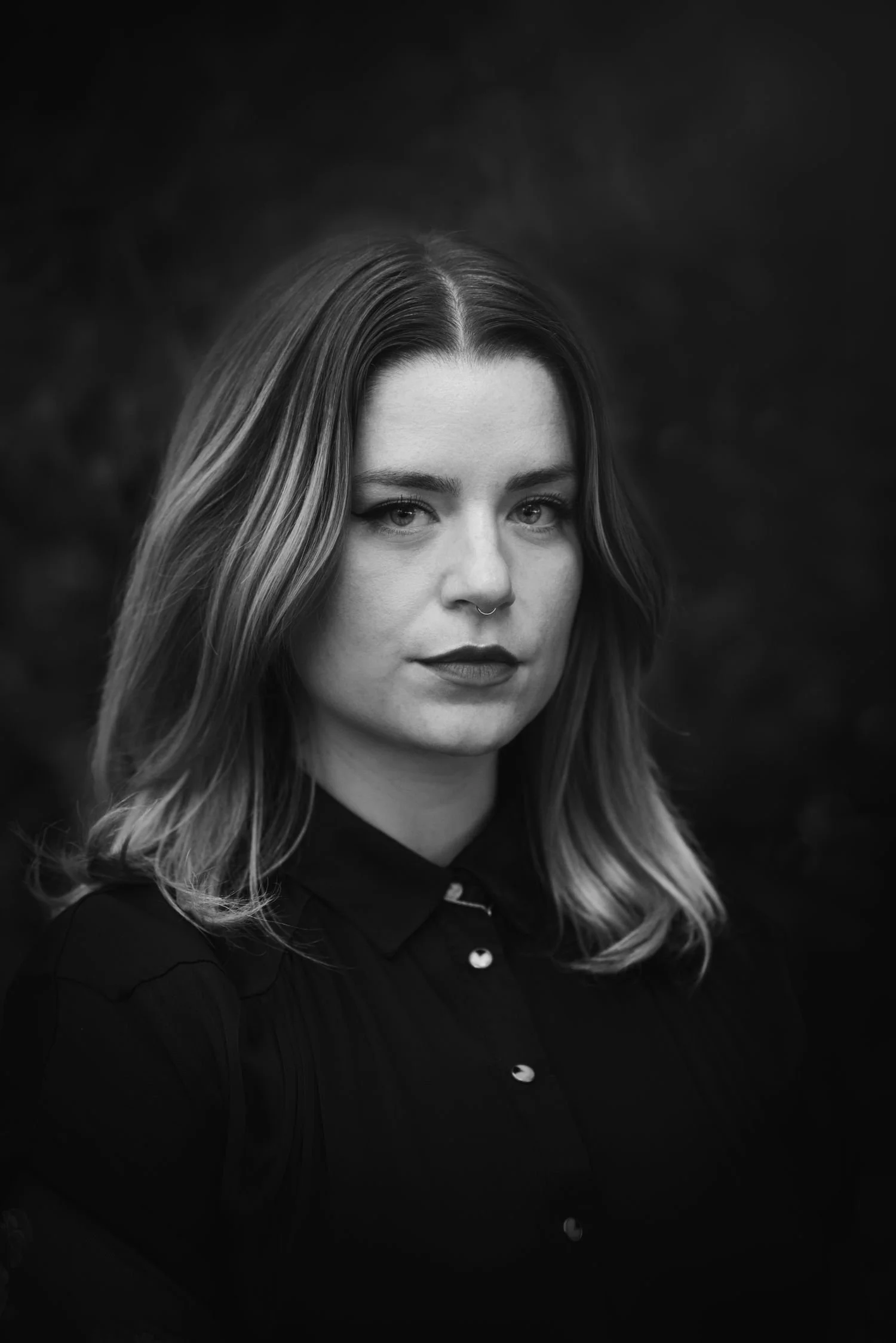 Portrait noir et blanc d'une femme aux cheveux longs ondulés, portant une chemise foncée, regardant vers l'objectif avec une expression sérieuse.