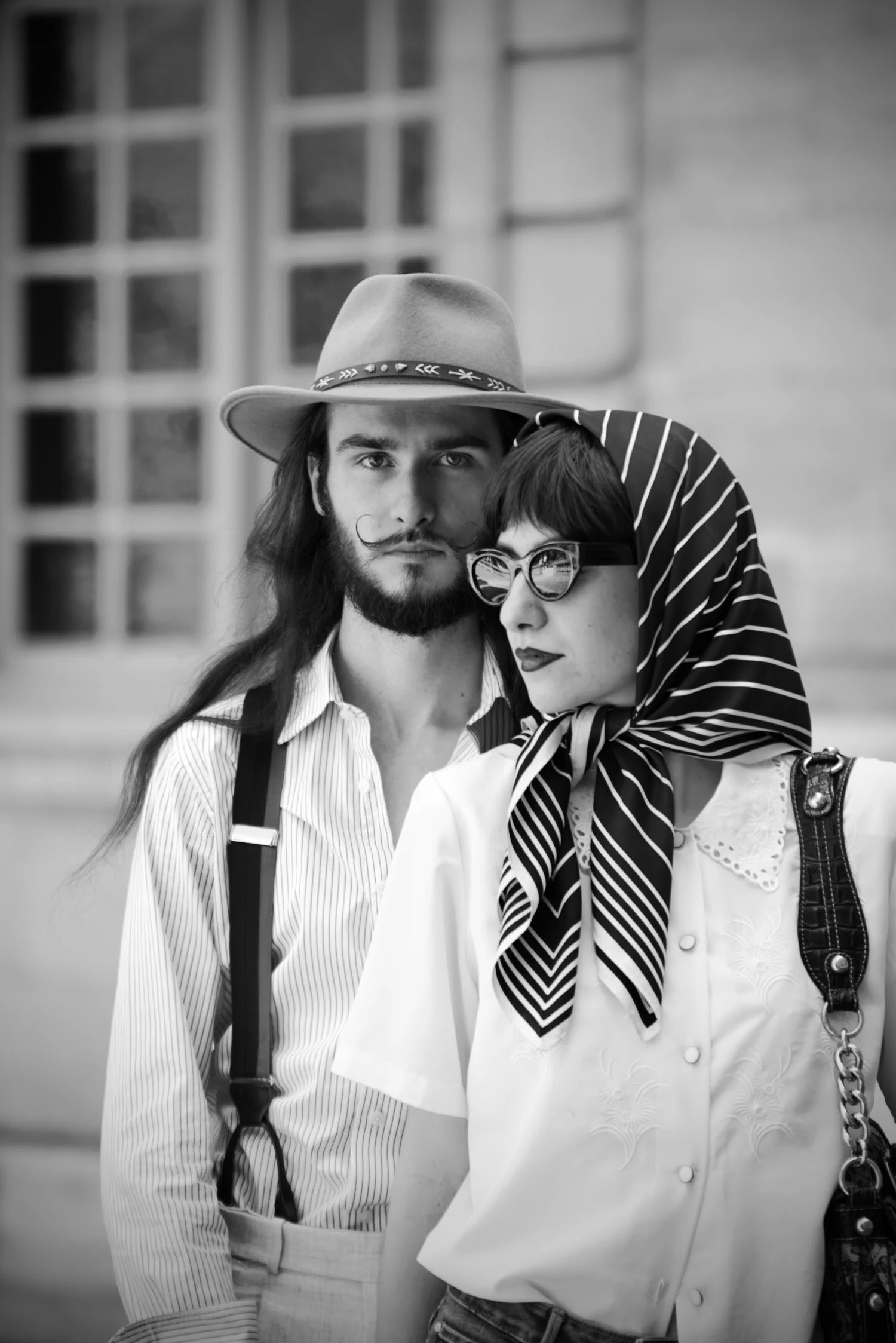 Deux personnes posent ensemble, un homme avec un chapeau et une femme avec des lunettes, dans un style vintage en noir et blanc.