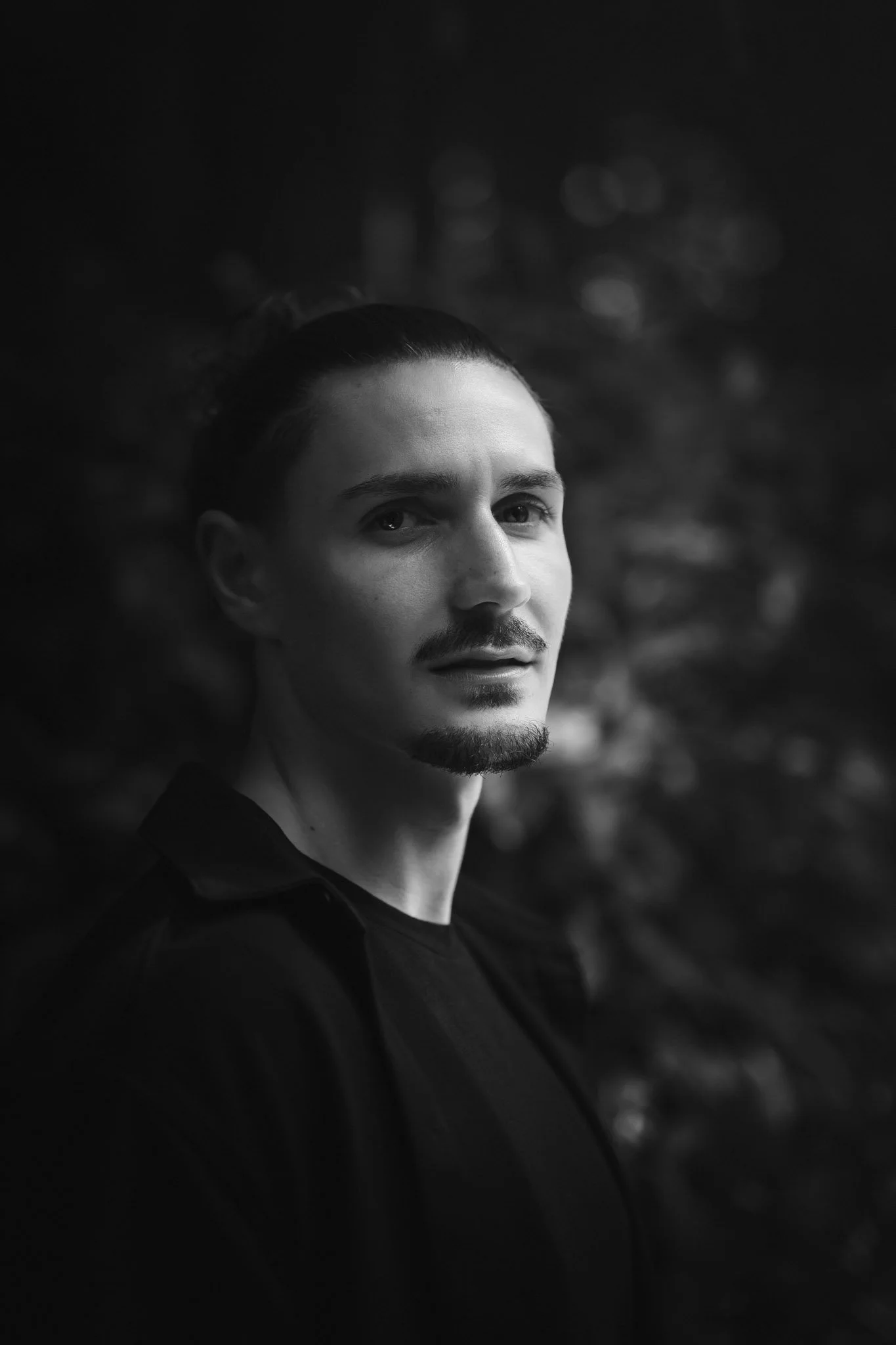 Portrait en noir et blanc d'un homme avec une barbe, portant une chemise noire, dans un décor naturel flou.