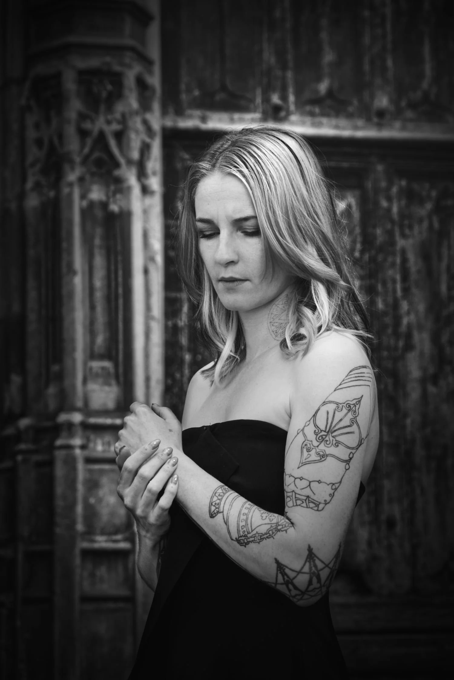 Une femme avec des tatouages, portant une robe noire, se tient dans une pièce sombre avec un décor en bois, regardant vers le bas avec une expression pensative.