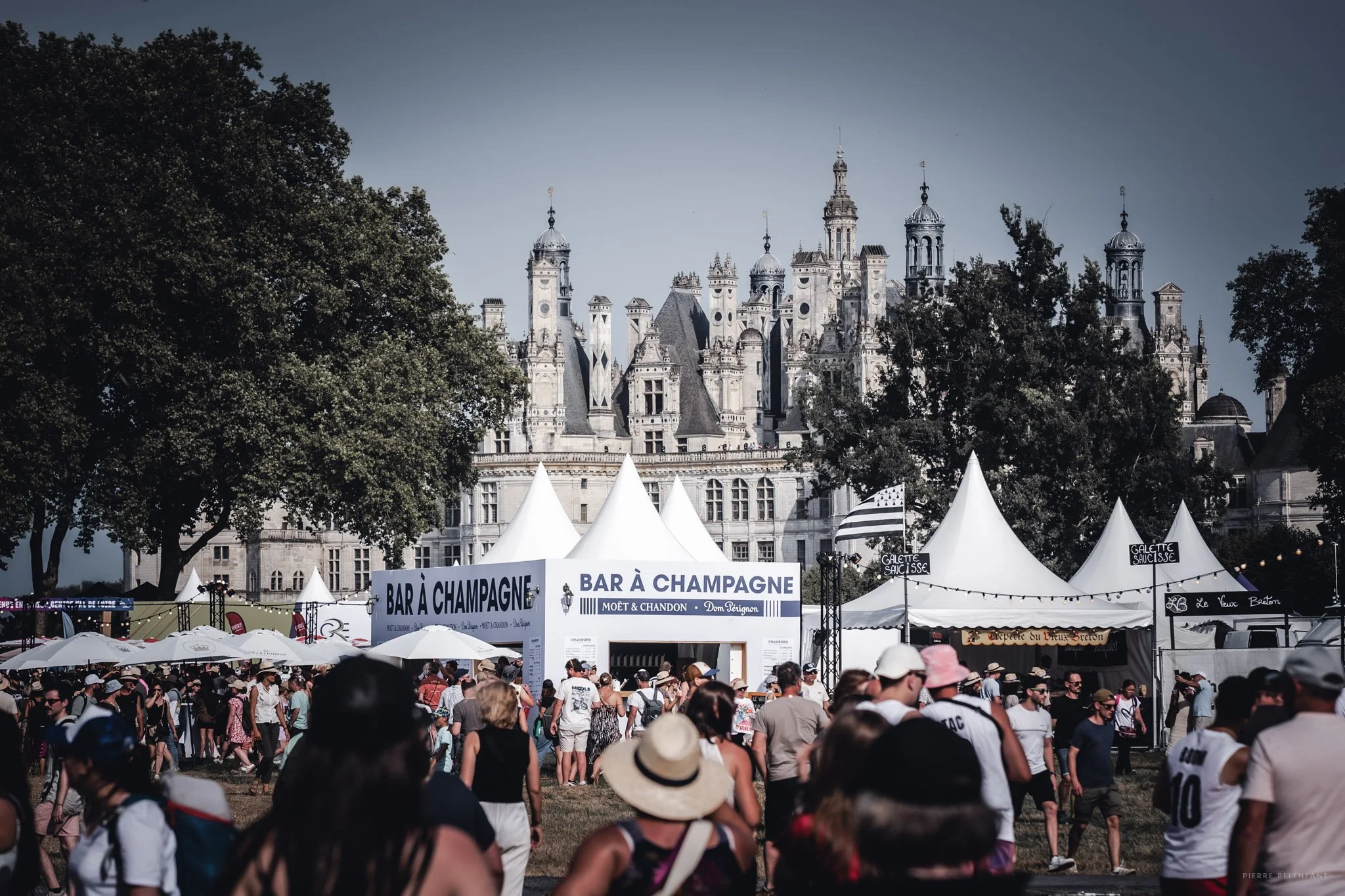 Fête en plein air avec des tentes blanches, des parasols et une foule de personnes sous un ciel teinté de bleu, avec le château de Chambord en arrière-plan.