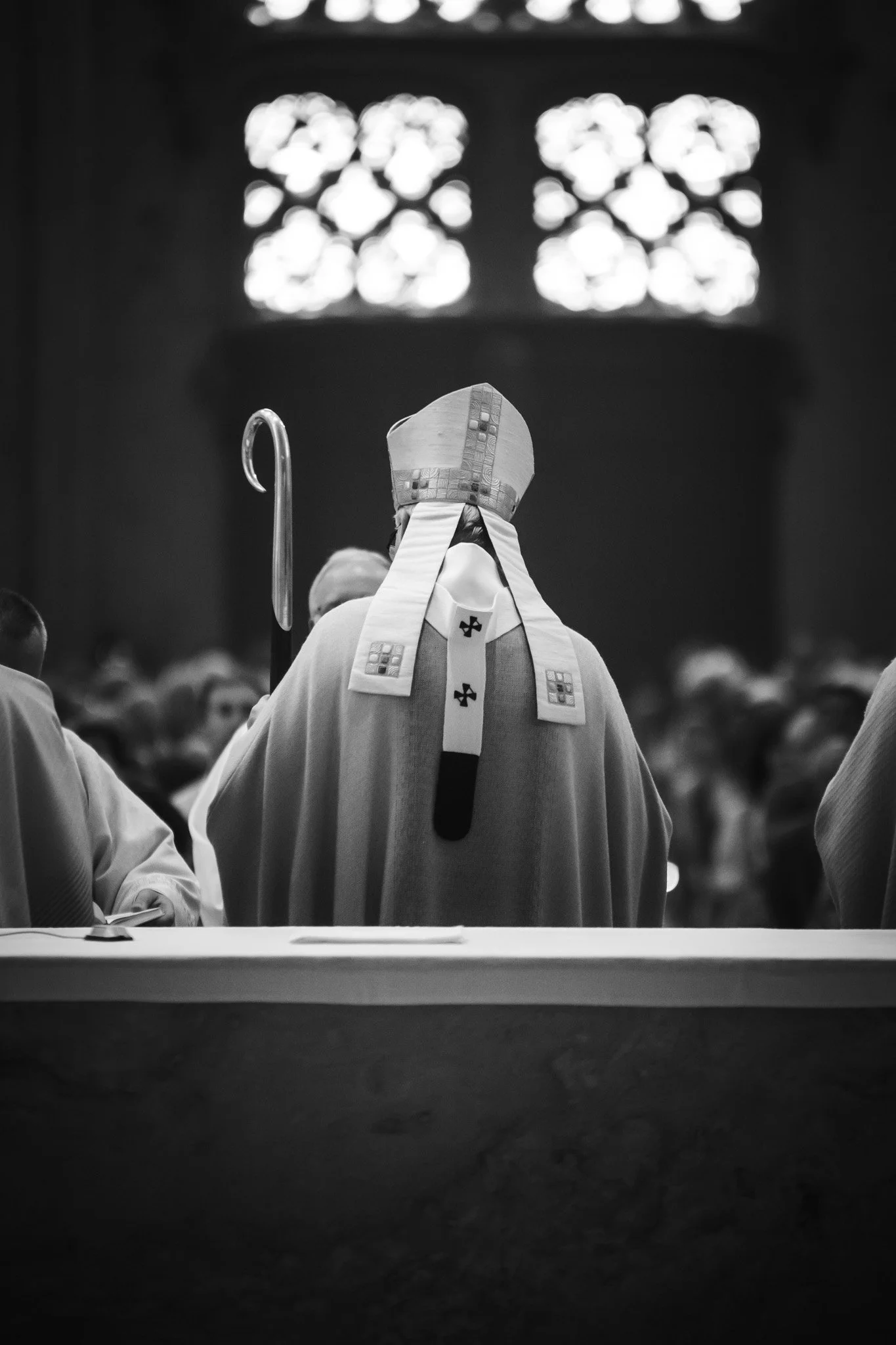 Prêtre en habits religieux, vu de dos, dans une église avec une grande fenêtre en vitrail en arrière-plan.