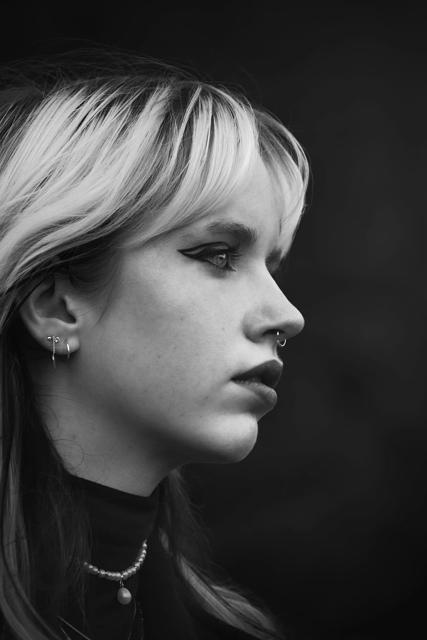 Un portrait en noir et blanc d'une jeune femme avec des cheveux longs et clairs, portant des boucles d'oreilles, un piercing nasal, un collier de perles et un maquillage avec du eyeliner marqué.