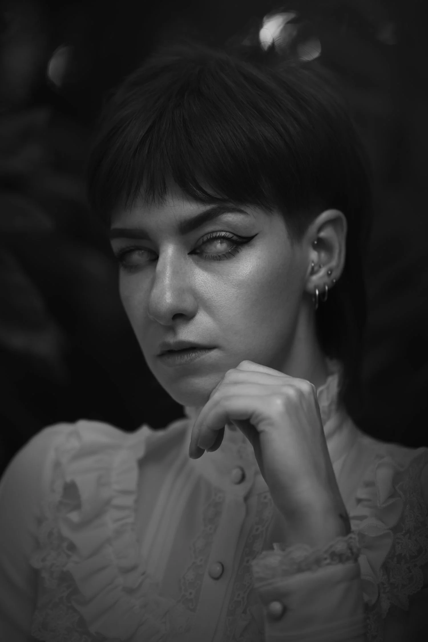 Portrait d'une femme au regard fixe, avec maquillage marqué, cheveux courts et vêtements à boutons et détails en dentelle, en noir et blanc.