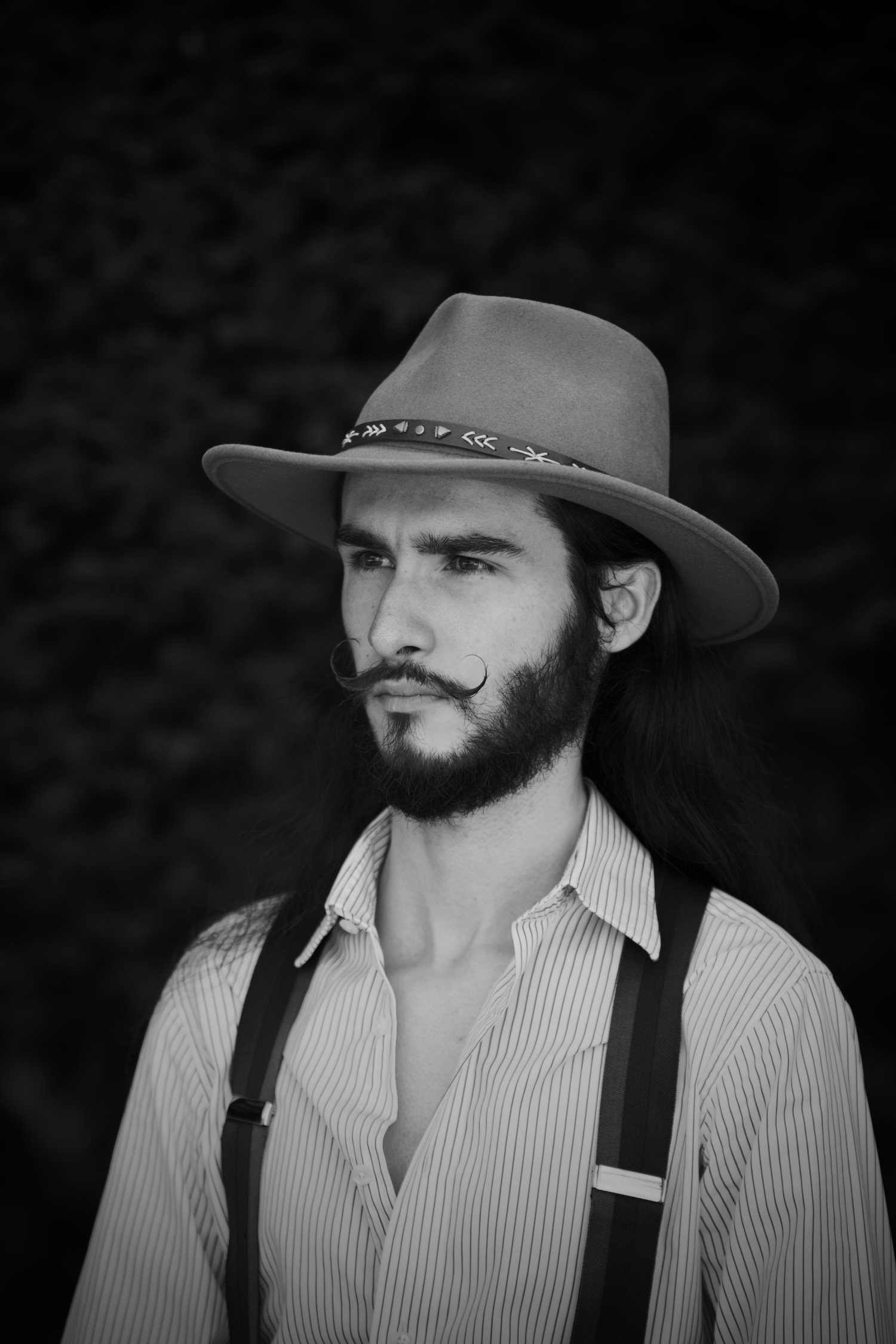 Portrait d'un homme avec une barbe et une moustache bouclée, portant un chapeau à large bord avec une bande décorative, une chemise à rayures verticales et des bretelles, regardant vers la gauche, en noir et blanc.