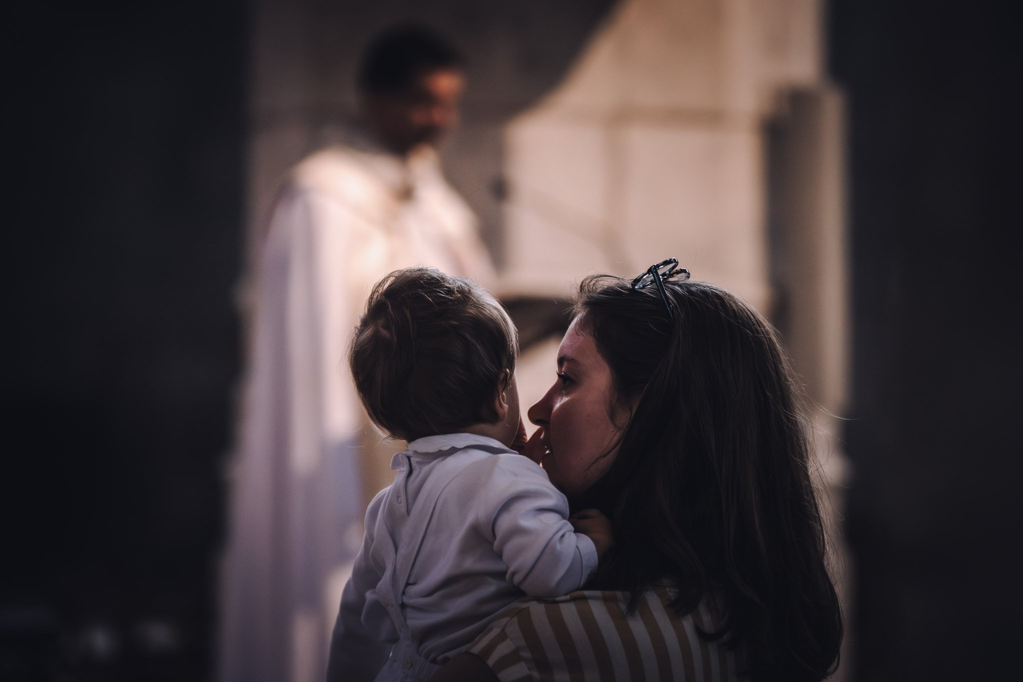 Une femme tient un jeune enfant dans ses bras, ils sont très proches et se regardent affectueusement, dans un lieu sombre.