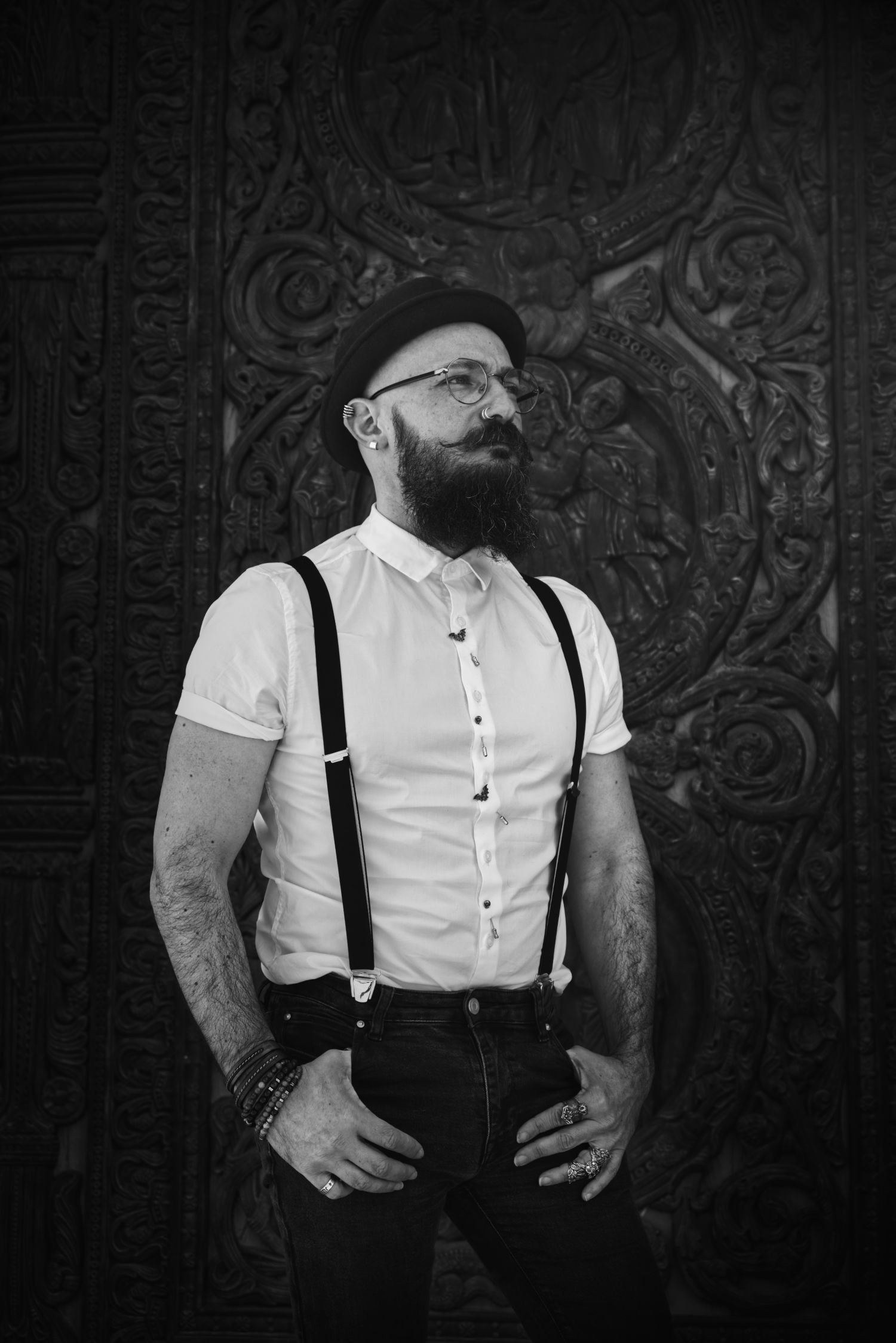 Homme avec un chapeau, lunettes, barbe et moustache, portant une chemise blanche à manches courtes, suspenders noirs, bracelets, bagues, posant devant une porte en bois ornée et sculptée.