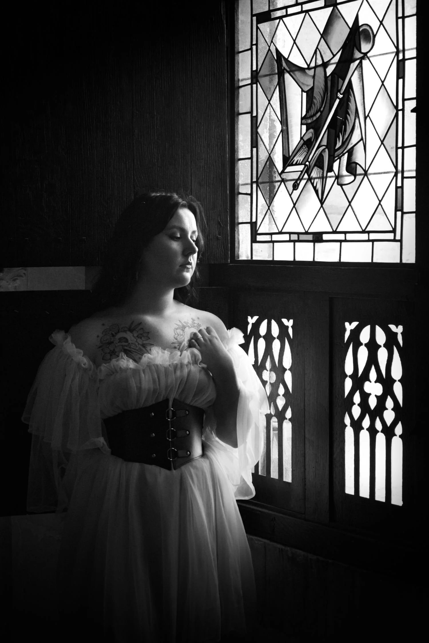 Une femme en robe blanche élégante se tient à côté d'une fenêtre en verre coloré avec des motifs religieux, dans une pièce sombre.