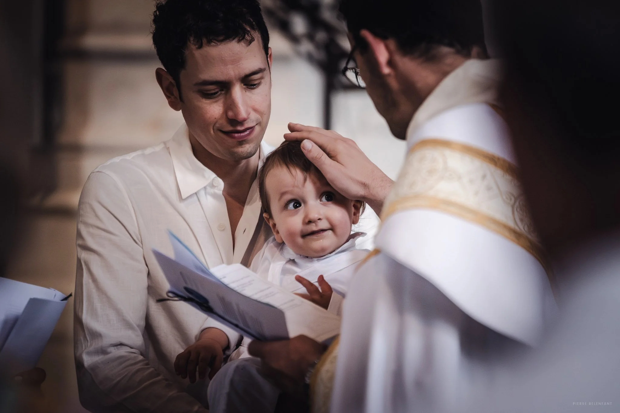 Un jeune homme, un bébé et un prêtre lors d'une cérémonie religieuse, probablement un baptême, dans une église.