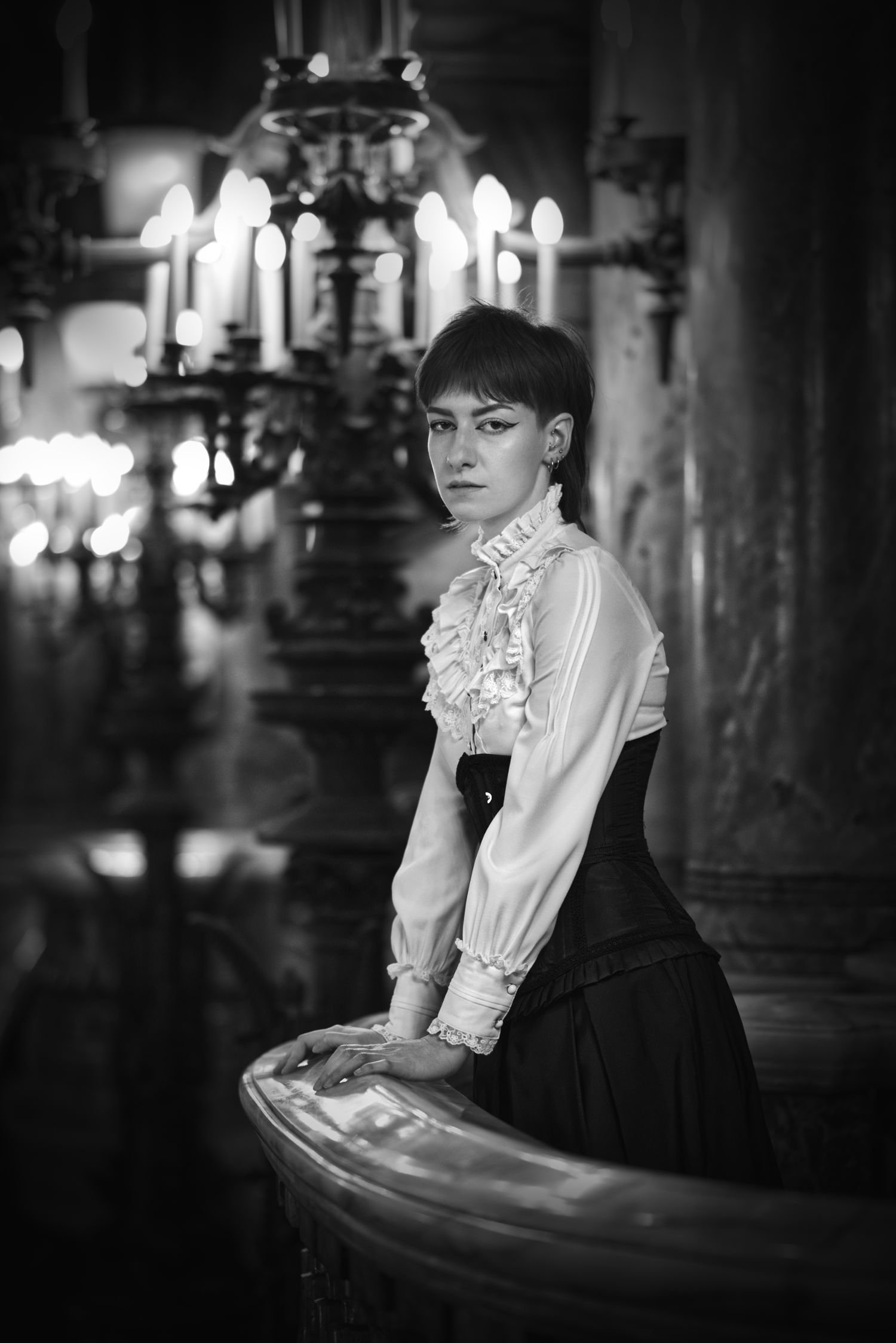 Une femme habillée d'une blouse à col froncé et d'une corset noire, posant sur une rampe dans une pièce ornée de candélabres allumés, en noir et blanc.