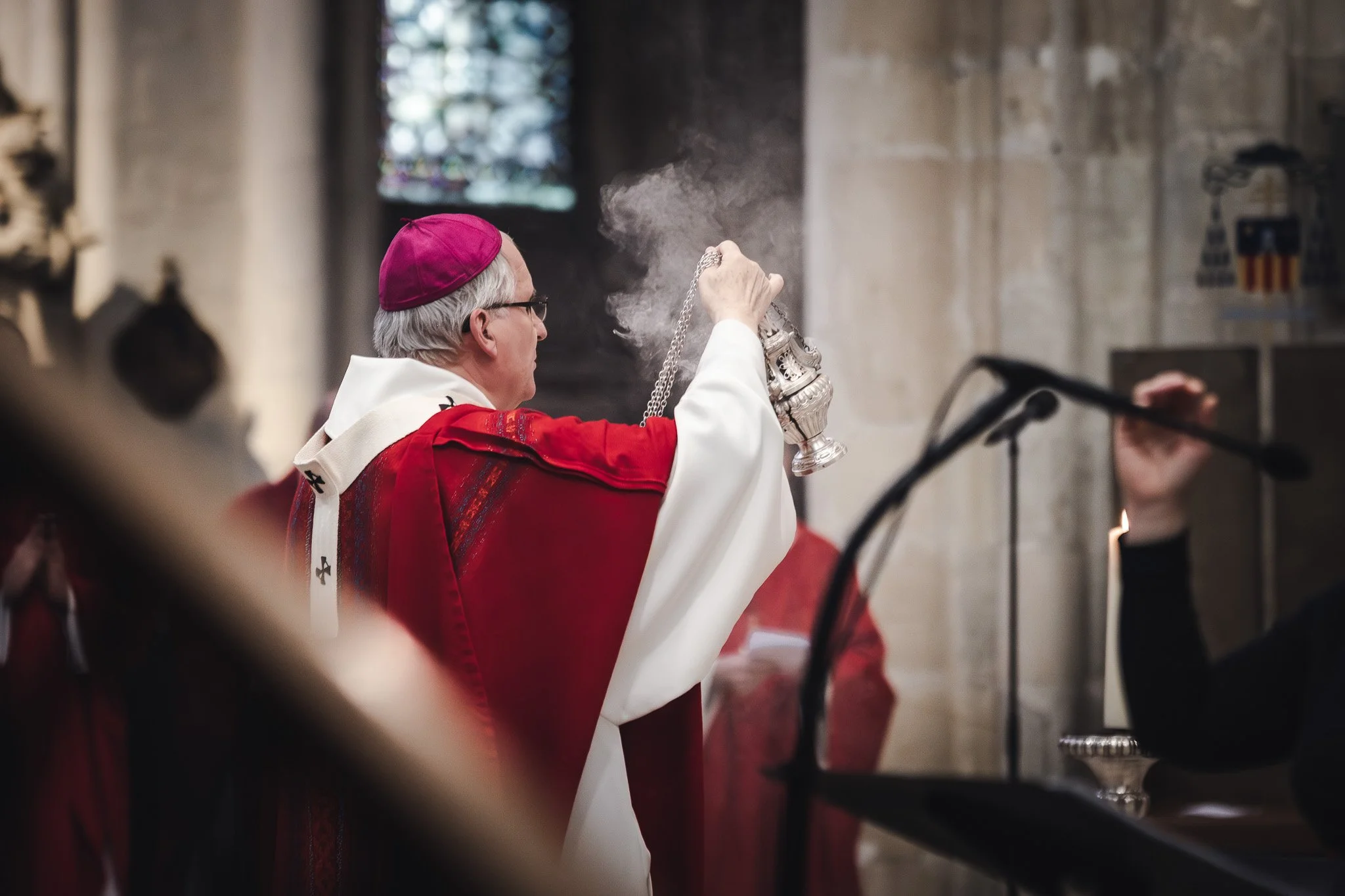 Un clerc catholique en robe rouge et blanche tenant un encensoir en fumée lors d'une cérémonie religieuse dans une église.