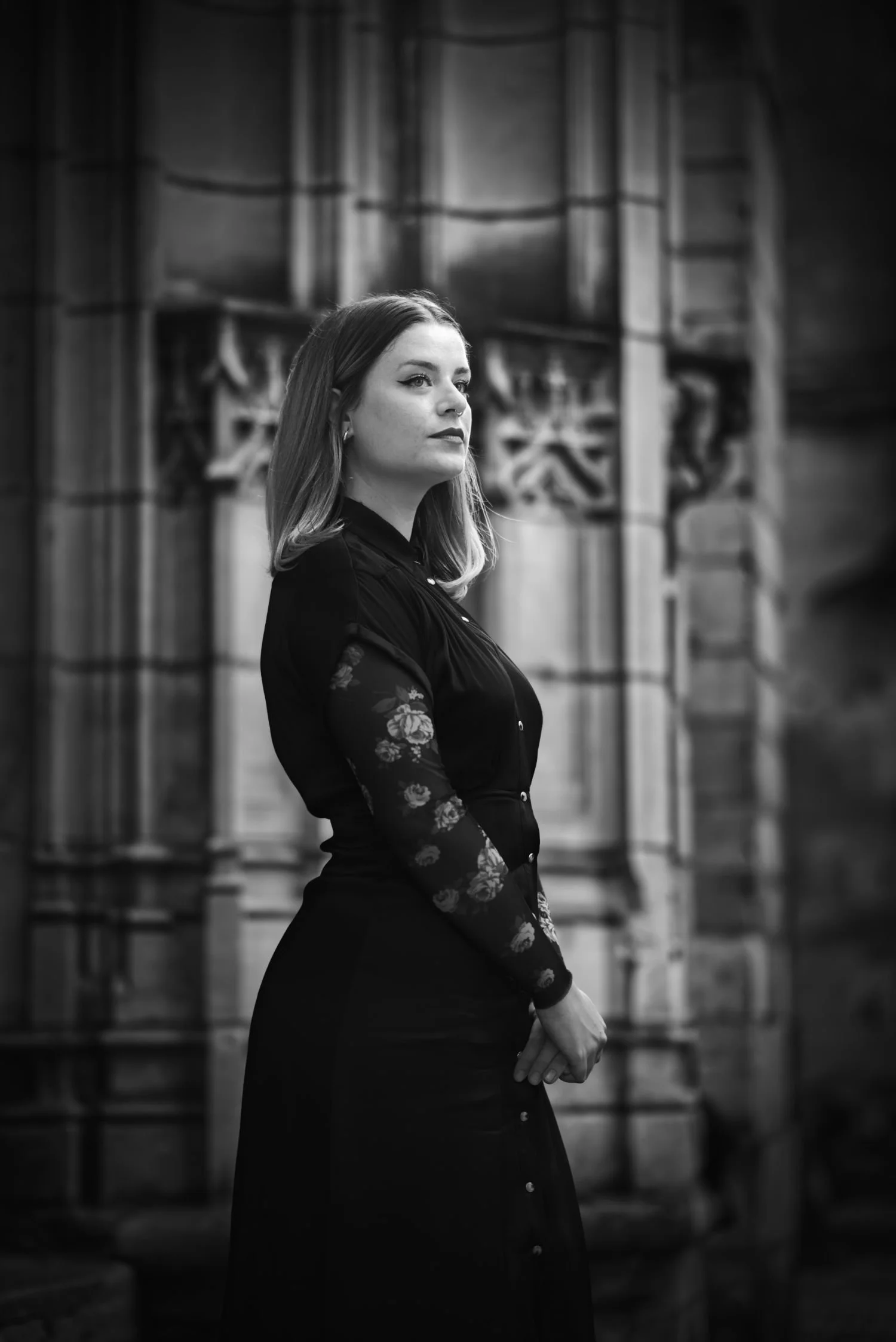 Une femme en portrait en noir et blanc, debout devant une architecture gothique avec des fenêtres en vitrail. Elle regarde de côté, portant une robe sombre avec des manches en dentelle à motifs floraux.
