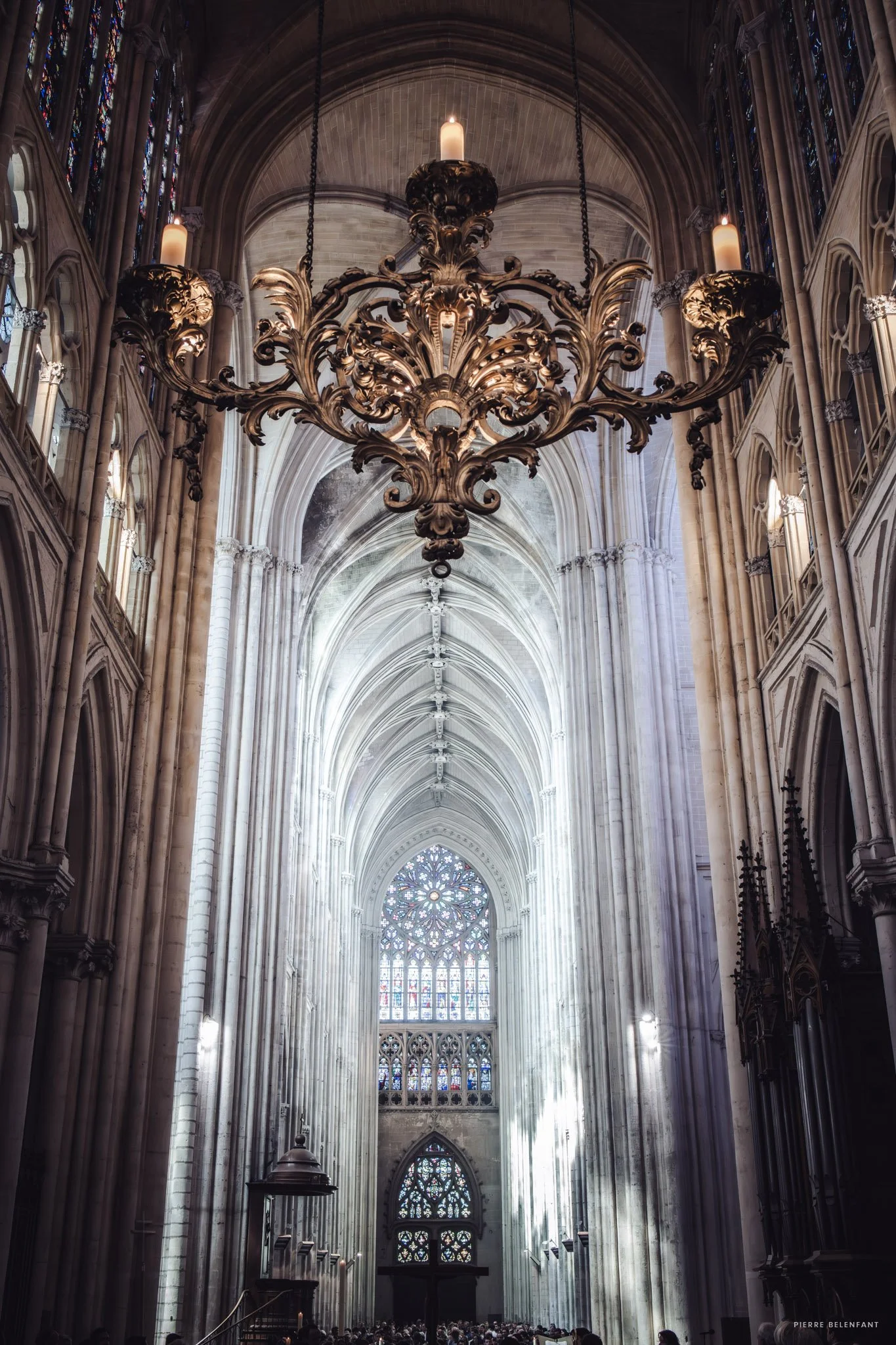 Intérieur d'une cathédrale gothique avec une grande rosace en vitrail au fond, un chandelier en or suspendu au plafond et des vitraux colorés sur les côtés.