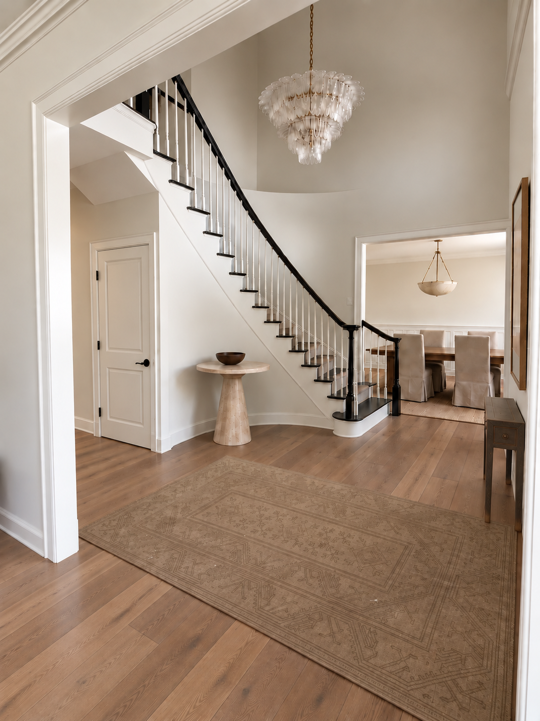 Interior view of a modern house entryway with a staircase featuring dark wood handrail, light-colored risers, and white spindles. There is a carpeted dining area in the background with a table, chairs, and a hanging light fixture. A beige area rug is on the wooden floor, and a small round table with a bowl on it is near the staircase.